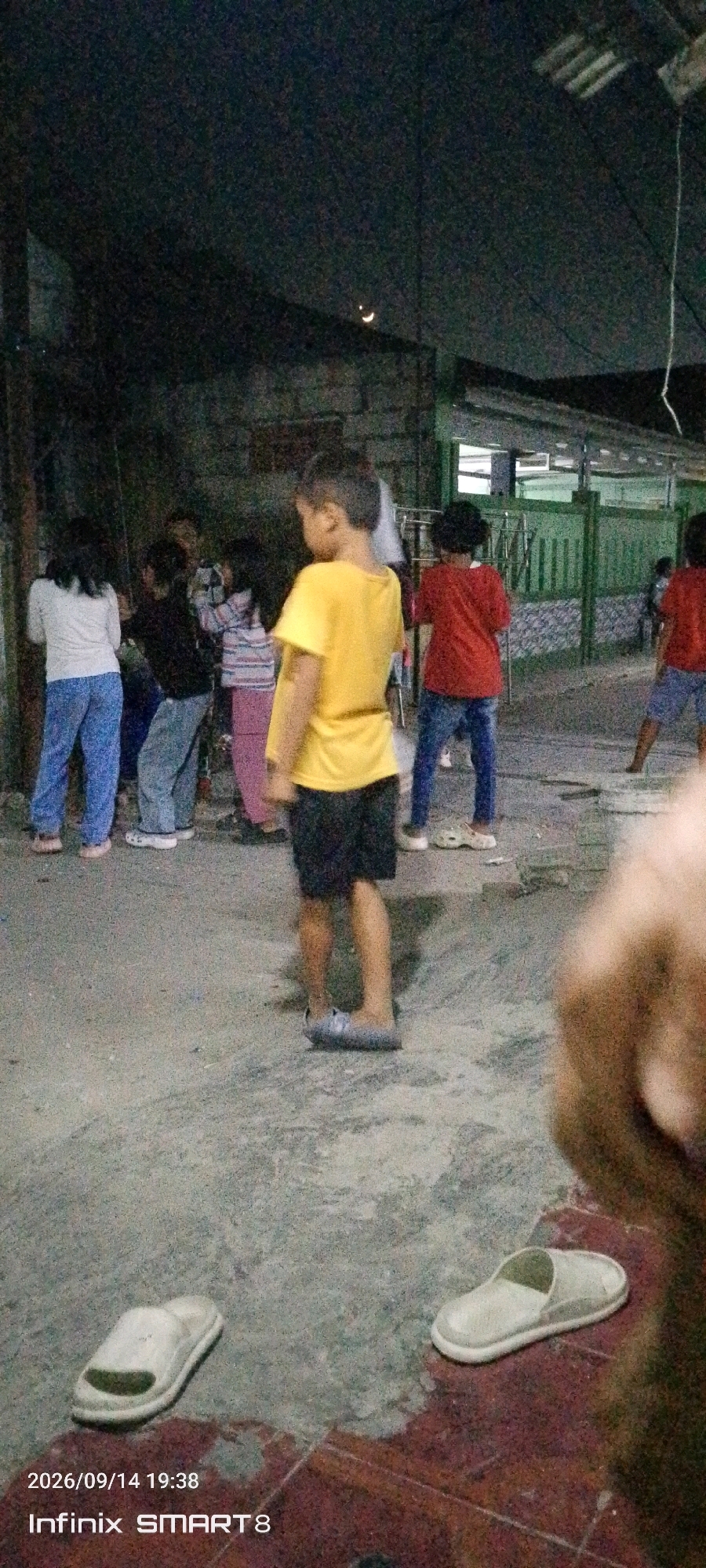In Serang, Indonesia, at an outdoor gathering possibly in a residential area or a community space, a group of children are visible. The scene is dimly lit, suggesting it is evening or night. A prominent crescent moon is visible in the dark sky. The ground appears to be paved with concrete and some reddish-brown tiles in the foreground. Two pairs of white slippers are placed on the ground.
In the center of the frame, a young boy wearing a yellow t-shirt and black shorts stands with his back to the camera, looking away. To his left, several other children are gathered, some appearing to be engaged in an activity. One girl on the far left wears a white long-sleeved shirt and blue pants. Next to her, a child in a dark top and grey pants stands. Further to the right, a child in a striped shirt and pink pants is visible.
To the right of the boy in yellow, another child in a red t-shirt and blue jeans is seen. In the background, a green fence and some structures suggest a built environment. The overall impression is of a casual, informal gathering of children outdoors in the evening. The timestamp at the bottom left corner indicates the image was taken on September 14, 2026, at 19:38.