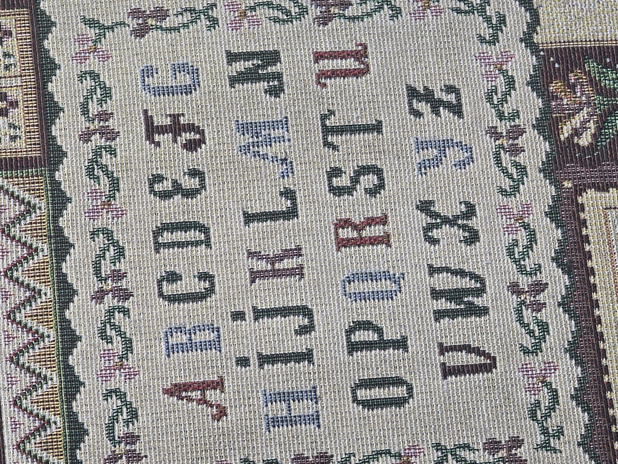 The image is a close-up, overhead view of a tapestry depicting the English alphabet, A through Z, in uppercase letters. The letters are arranged in four rows, with the first row containing A through G, the second row H through N, the third row O through U, and the fourth row V through Z. The tapestry has decorative borders above and below the alphabet, featuring floral patterns and geometric designs in various colors. The tapestry is made of woven fabric, likely a needlepoint or cross-stitch, with the letters and designs formed by stitching. There are no people or other objects visible in the image, and no information is provided about its location in Ulan-Ude, Russia.