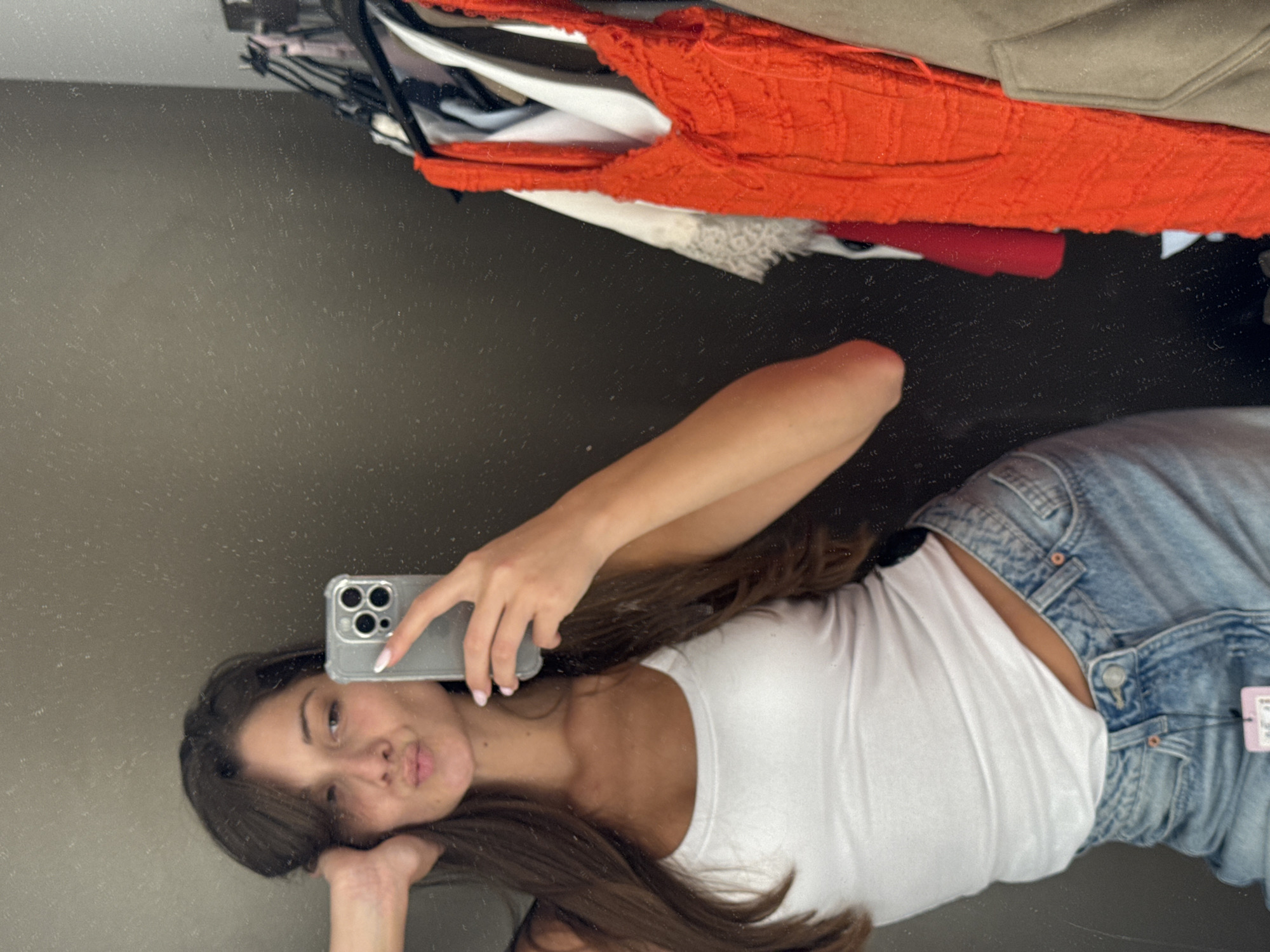 A young woman with long brown hair is taking a selfie in a fitting room. She is wearing a white tank top and light blue jeans. She is puckering her lips and holding a smartphone in her right hand, which is positioned to take the photo. Her left arm is bent and her hand is resting on her head. In the background, on a clothing rack, are several garments, including a bright orange textured top. The fitting room walls are a dark, speckled gray. No specific location in Burgas, Bulgaria is evident from the image.