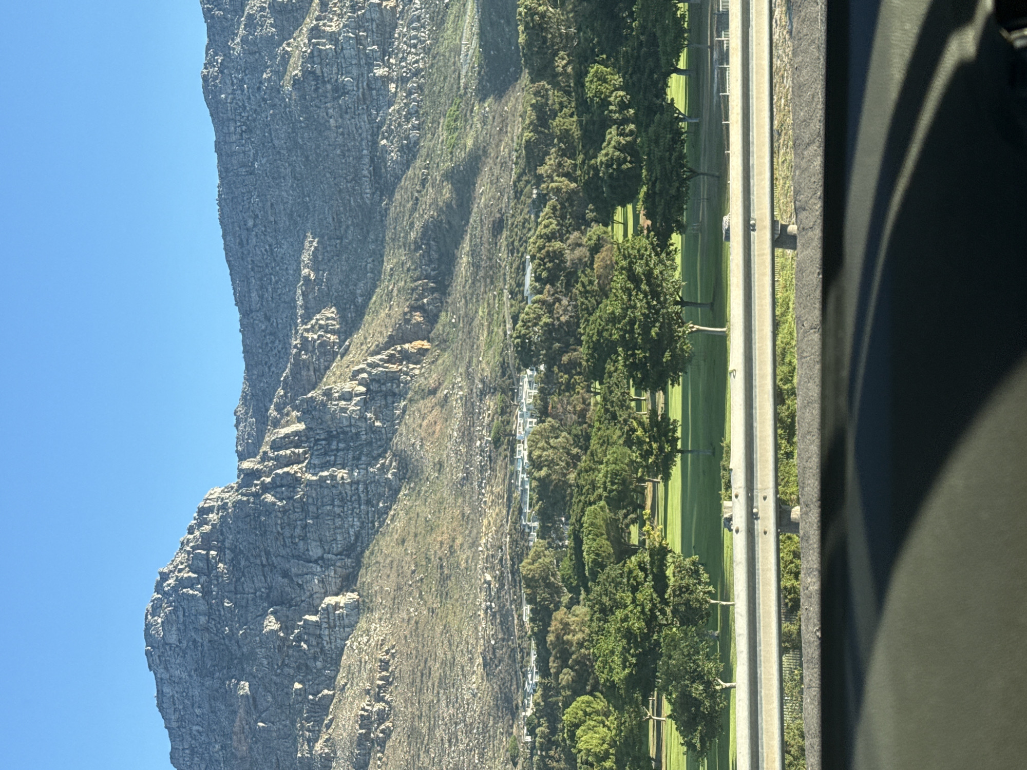 The image shows a view of Table Mountain in Cape Town, South Africa. The mountain, a massive rock formation, occupies the left side of the frame under a clear blue sky. To the right of the mountain, there is a strip of land, featuring vegetation, buildings, and a multi-lane highway. Cars are visible on the road. A portion of a vehicle interior intrudes into the right edge of the frame, indicating the image was taken from inside a car. The orientation of the photo is tilted to the left.