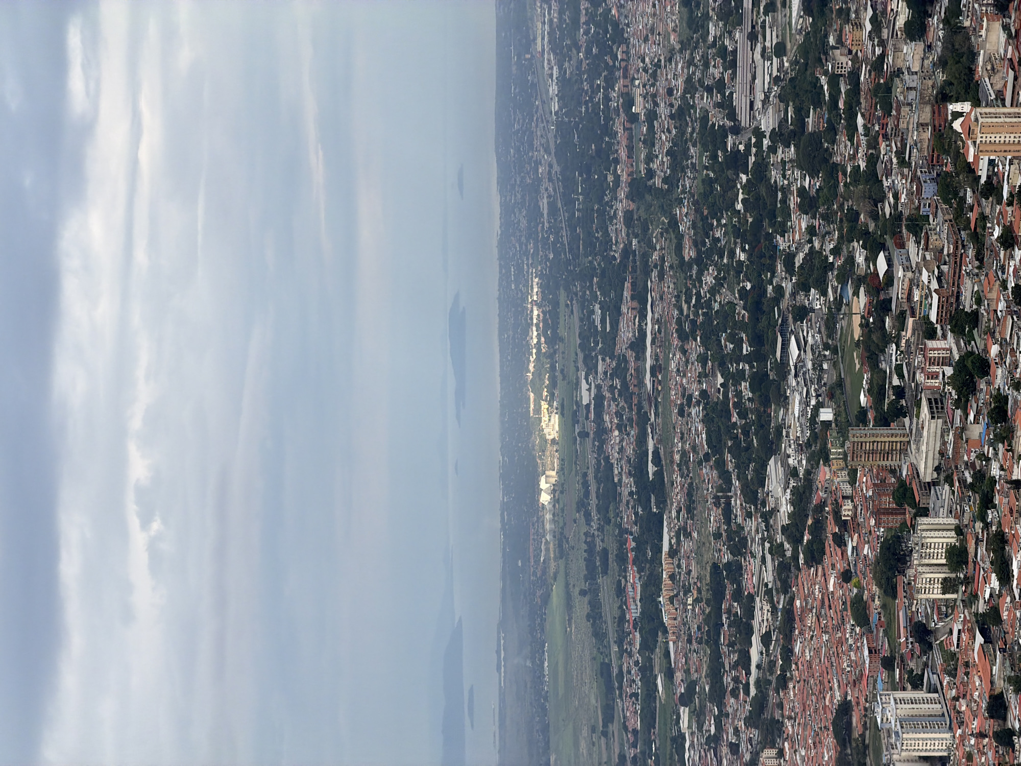 This is an aerial, high-angle, eye-level shot of a dense urban landscape, characteristic of a city in Venezuela, possibly El Cambur, given the context provided. The image captures a sprawling city with a mix of residential and commercial buildings. In the foreground, a dense collection of houses with terracotta roofs dominates, interspersed with taller, modern buildings, including high-rises. The mid-ground reveals a more varied urban fabric with commercial structures, industrial-looking buildings with large roofs, and green spaces. A highway or major road is visible winding through the city. In the far distance, a body of water, likely a lake or bay, is visible, with islands dotting its surface. The horizon is marked by hazy hills or mountains. The sky is overcast with layers of clouds, suggesting a subdued or perhaps late afternoon light. The scene is devoid of any visible people or specific actions, presenting a static view of the city's architecture and layout. The overall impression is one of a heavily populated urban area with significant development.