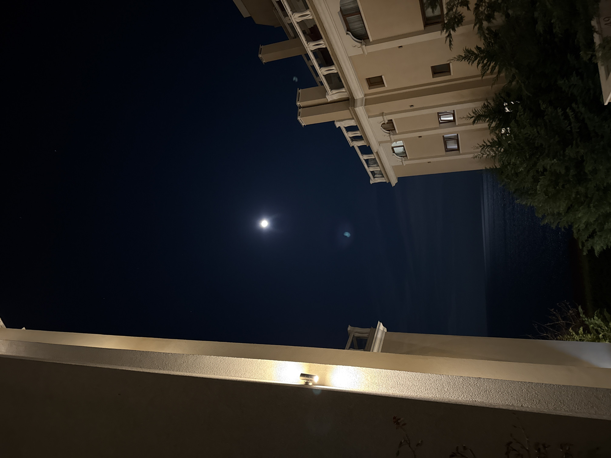 The image captures a night scene in what appears to be Varna, Bulgaria, recognizable by the architectural style and coastal proximity. The viewpoint is looking upwards, encompassing a dark, clear night sky illuminated by the moon. A tall, beige-colored building with multiple floors and balconies dominates the upper portion of the frame. It exhibits architectural details consistent with buildings found in Varna. A dark body of water, presumably the Black Sea, can be seen adjacent to the building. Dense, dark green foliage is present on the right side of the image, potentially trees or bushes. In the foreground, a section of a wall or balcony railing is visible, illuminated by a small, built-in light fixture. The scene lacks people or active events.