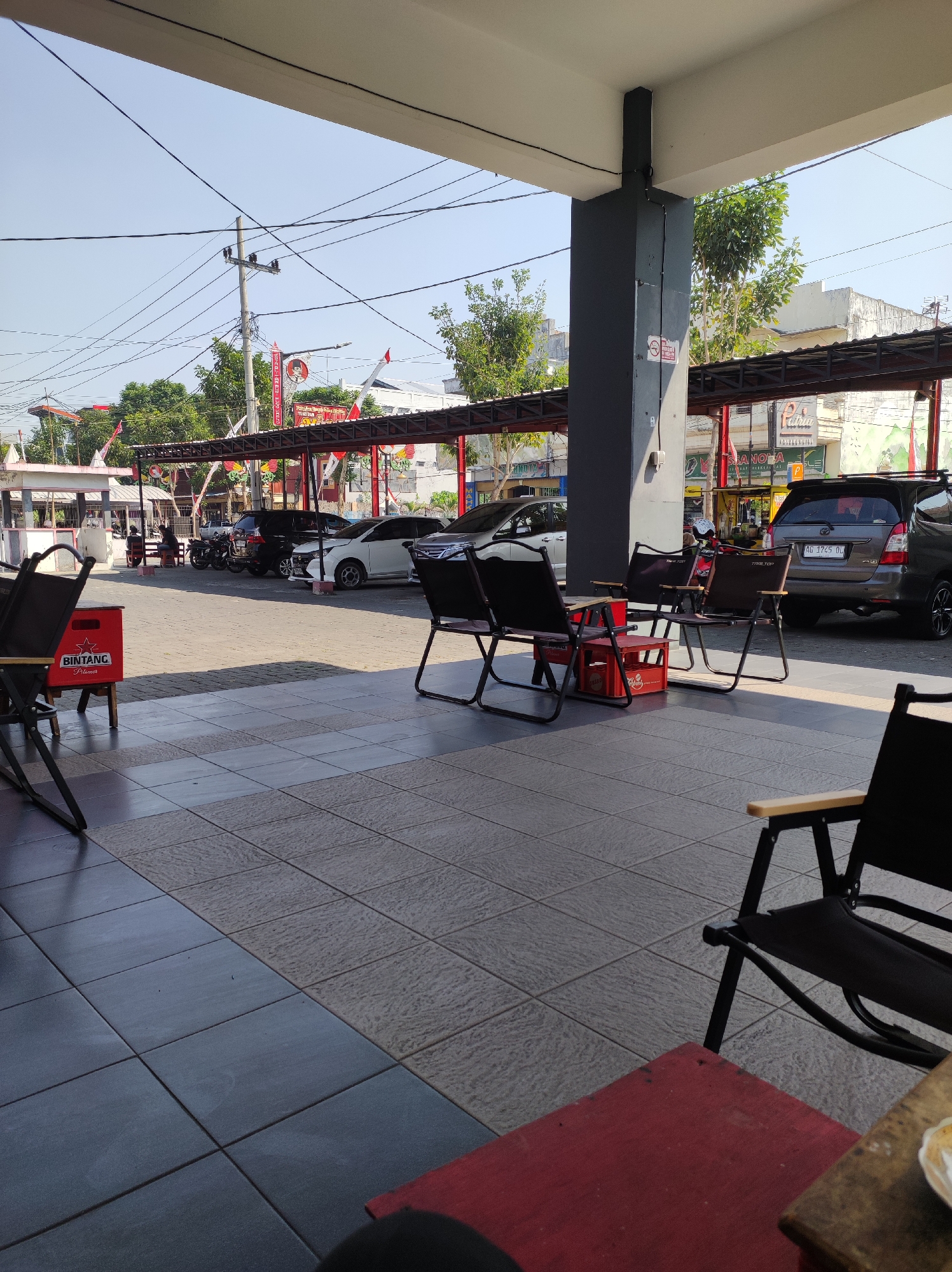 The image depicts an outdoor seating area with several cars parked in the background under a covered structure. The foreground shows a tiled floor and several empty folding chairs, some with a red crate labeled "BINTANG" underneath. Beyond the chairs, a row of cars, including a grey SUV with license plate AG 1745 DL, and a white sedan, are parked. A motorcycle is also visible. The background features buildings, trees, and power lines against a clear sky. The scene suggests a casual outdoor space, possibly a cafe or waiting area, in Blitar, Indonesia, as indicated by the presence of Indonesian license plates and general architecture.
