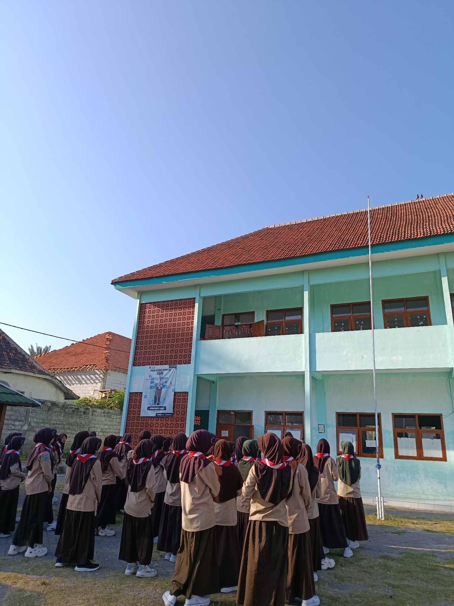In Ramban Wetan, Indonesia, a group of young individuals, likely students, are gathered outdoors in front of a light blue building. They are all wearing uniforms consisting of brown skirts, light brown shirts, and dark brown headscarves, with red and white sashes across their chests, suggesting a scouting or similar organization. The students are facing away from the camera, towards the building and a flagpole. The building has multiple windows and a red tiled roof. A large banner is affixed to the building's facade. The ground is a mix of paved areas and grass. The sky is clear and blue, indicating a sunny day.