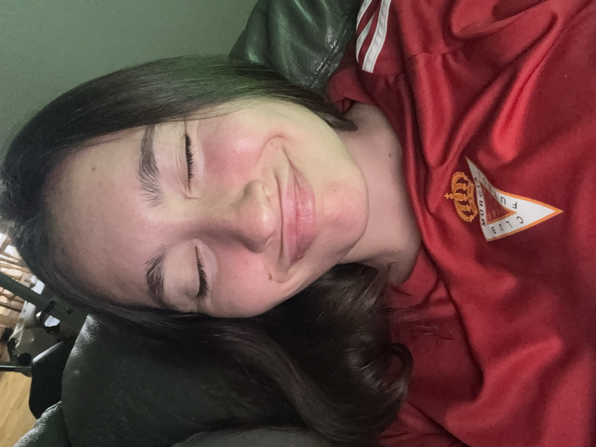 A young woman with closed eyes and a puckered smile poses for a selfie. She has long, dark brown hair and is wearing a red t-shirt with white stripes on the sleeves. A logo, partially visible, appears on the shirt. The background is blurred, but suggests an indoor setting with green walls and dark furniture. The image is taken at a close-up, head-and-shoulders perspective.