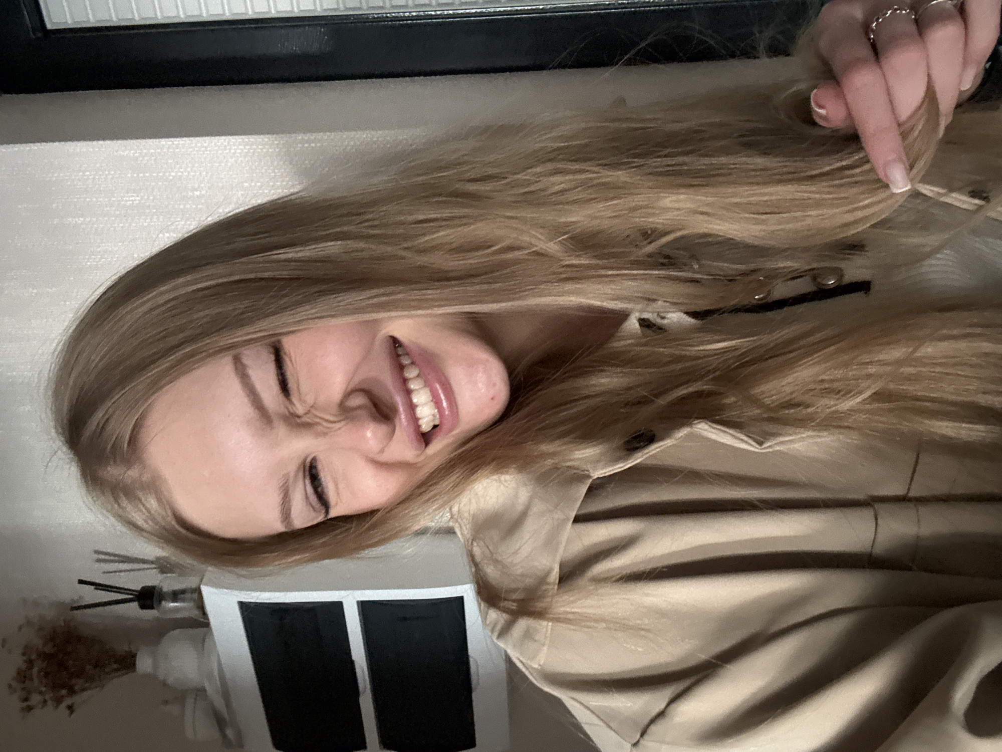 A young woman with long blonde hair is winking and grinning, showing her teeth, in a selfie. She is wearing a beige jacket over a light-colored shirt. Her right hand is touching her hair near her chin. She is wearing two rings on her right ring finger. In the background, to the left, there is a white shelf with decorative items, including a vase with dried flowers and two diffusers. To the right, a textured wall is visible, and a black-framed window with blinds. The lighting is dim. There is no indication of the city of Perm, Russia, in the image.