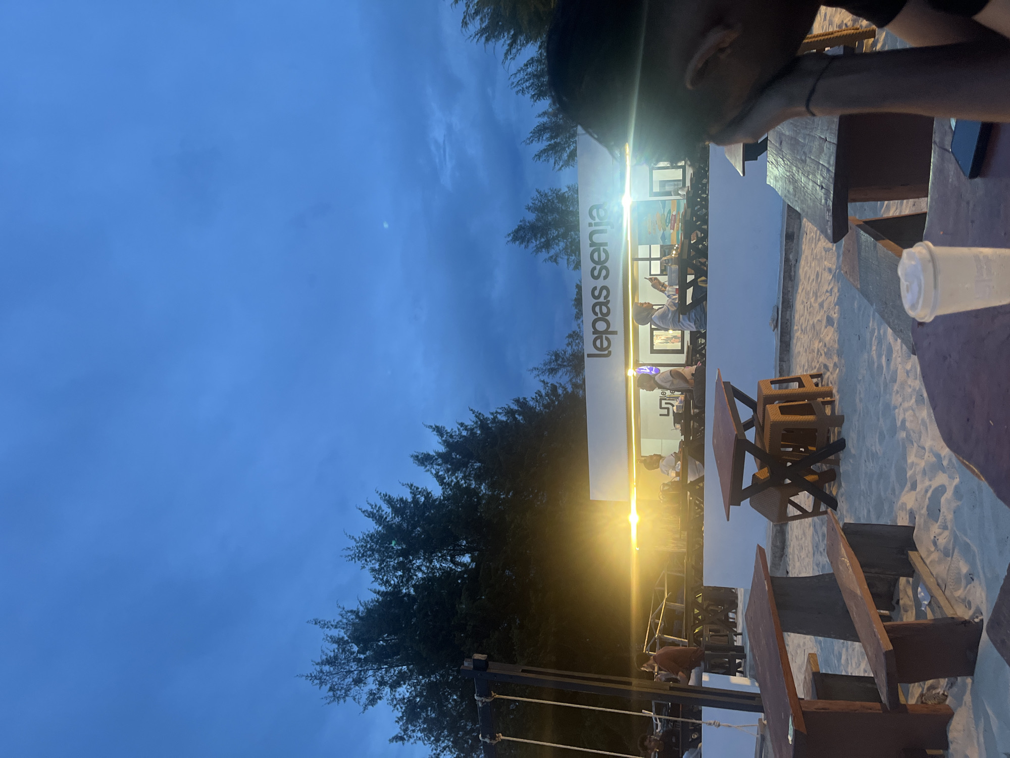 The image shows a beachfront cafe called "Lepas Senja" in Monikeun, Indonesia. It is dusk, and the sky is a deep blue with some clouds. The cafe has outdoor seating on a sandy beach. There are several wooden tables and benches set up, some with chairs. In the background, there is a building with bright lights, presumably part of the cafe. People are seated at tables inside and outside the cafe. A person's head and shoulder are visible in the foreground, obscuring part of the scene. Trees are visible behind the cafe.