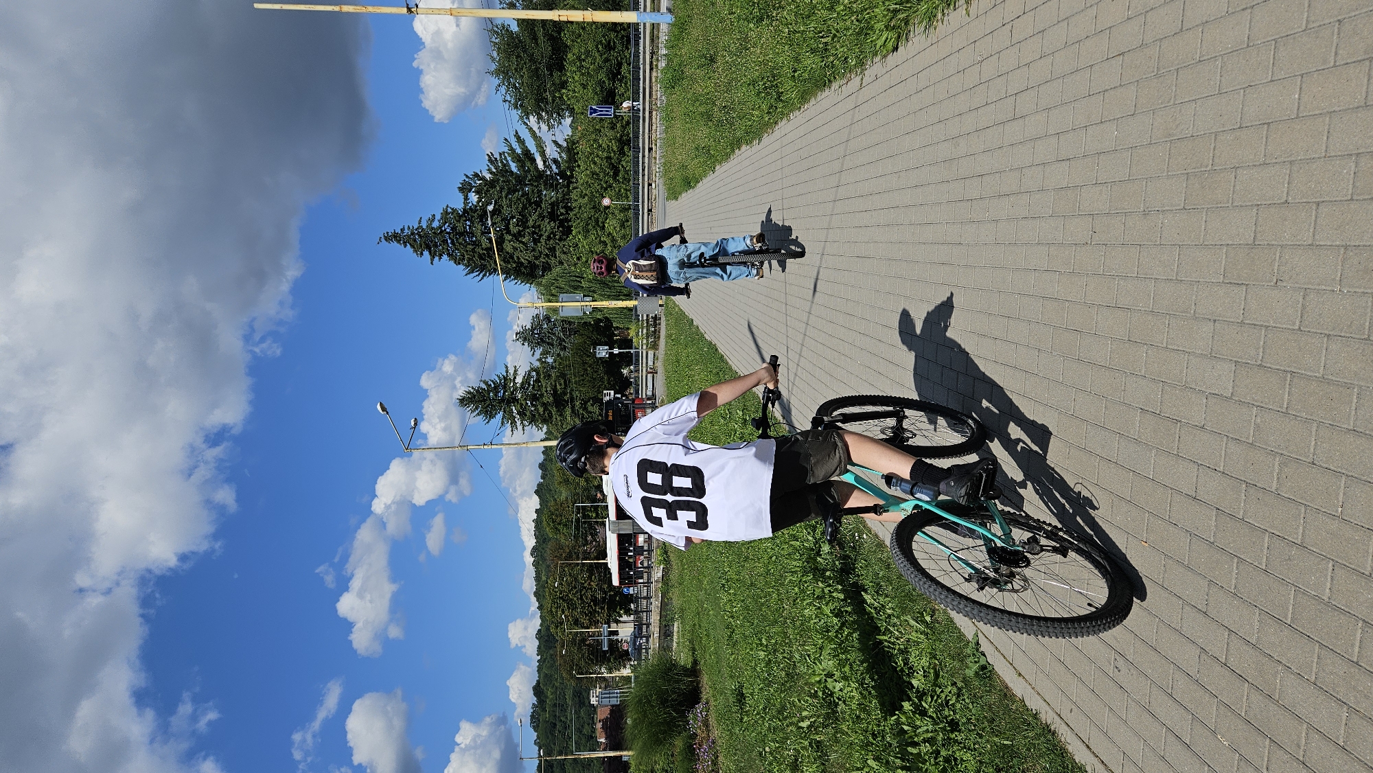 The image depicts a sunny day in Prešov, Slovakia, with two individuals cycling along a paved pedestrian path. The path is bordered by a grassy area on the left and trees on the right. The sky is a vibrant blue with scattered white and gray clouds. In the background, there are buildings, trees, and a road with a bus. The foreground focuses on the cyclists. The person in the front, facing away from the camera, is wearing a white t-shirt with the number "38" in black on the back, a black helmet, and shorts. They are riding a teal-colored bicycle. The second cyclist, further ahead, is also facing away and wearing a blue top, jeans, a helmet, and a backpack. They are riding a bicycle with a black frame. The scene suggests a leisurely outdoor activity in an urban or suburban setting.