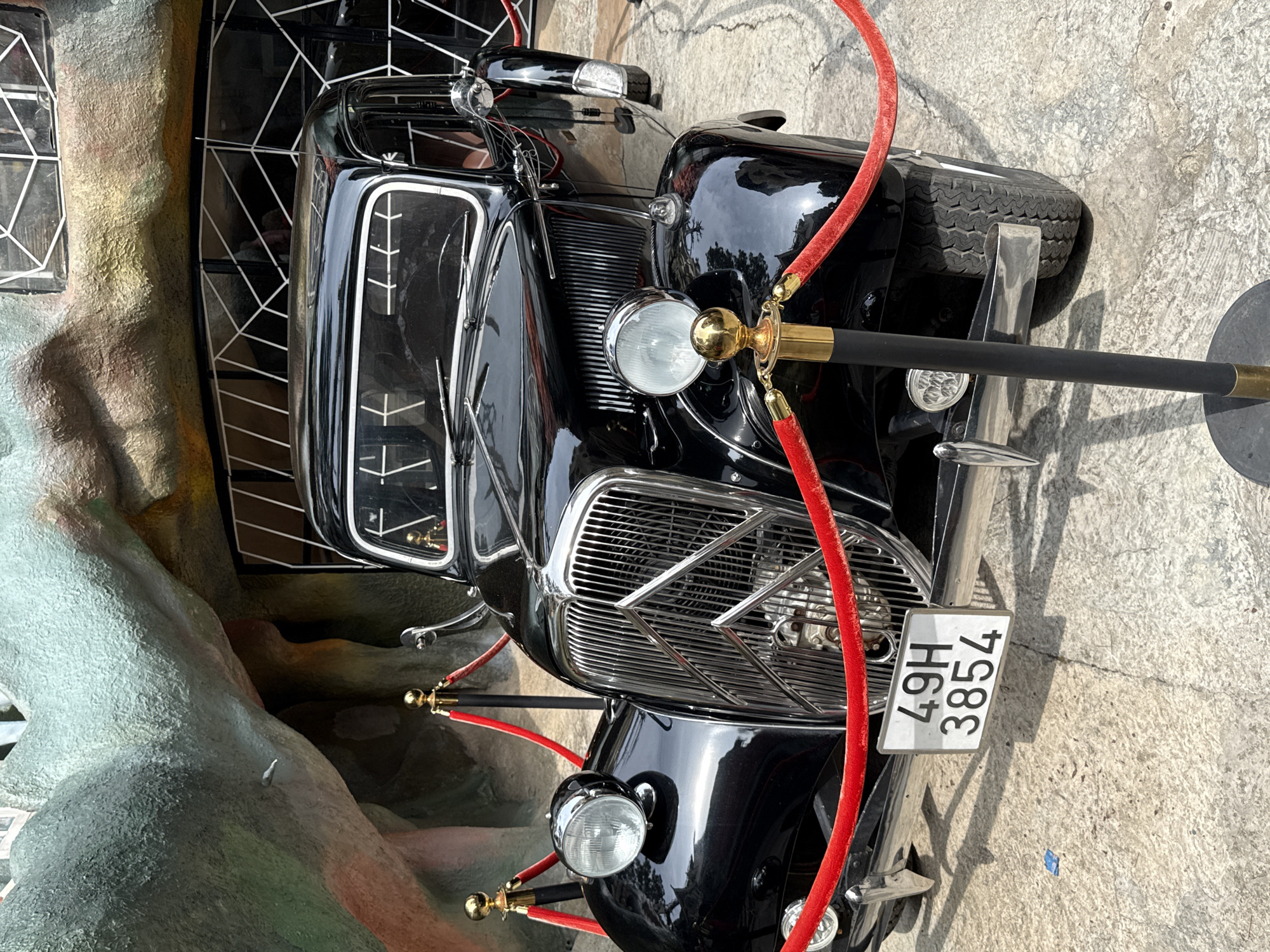This image displays a vintage black car, likely a Citroën Traction Avant, on display outdoors in Da Lat, Vietnam. The car is pristinely maintained with a glossy black finish and chrome accents, including its prominent front grille and round headlights. A red velvet rope, held by ornate gold stanchions, forms a barrier in front of the vehicle, indicating it is an exhibit.
Behind the car, an unusual, organic-shaped structure resembling rock or concrete formations is visible. This structure is textured and features windows with distinctive white, angular grid patterns, suggesting a unique architectural style. The colors of the structure range from light gray and green to earthy tones.
The car is positioned on a light gray concrete surface, which shows some minor cracks and shadows consistent with daytime lighting on a clear day. A spare tire is mounted at the rear of the vehicle. The front license plate clearly displays the text "49H 3854". No individuals are present in the scene, allowing the vintage car and its distinctive surroundings to be the central focus. The setting suggests the car is part of a tourist attraction or a themed display within Da Lat, Vietnam.