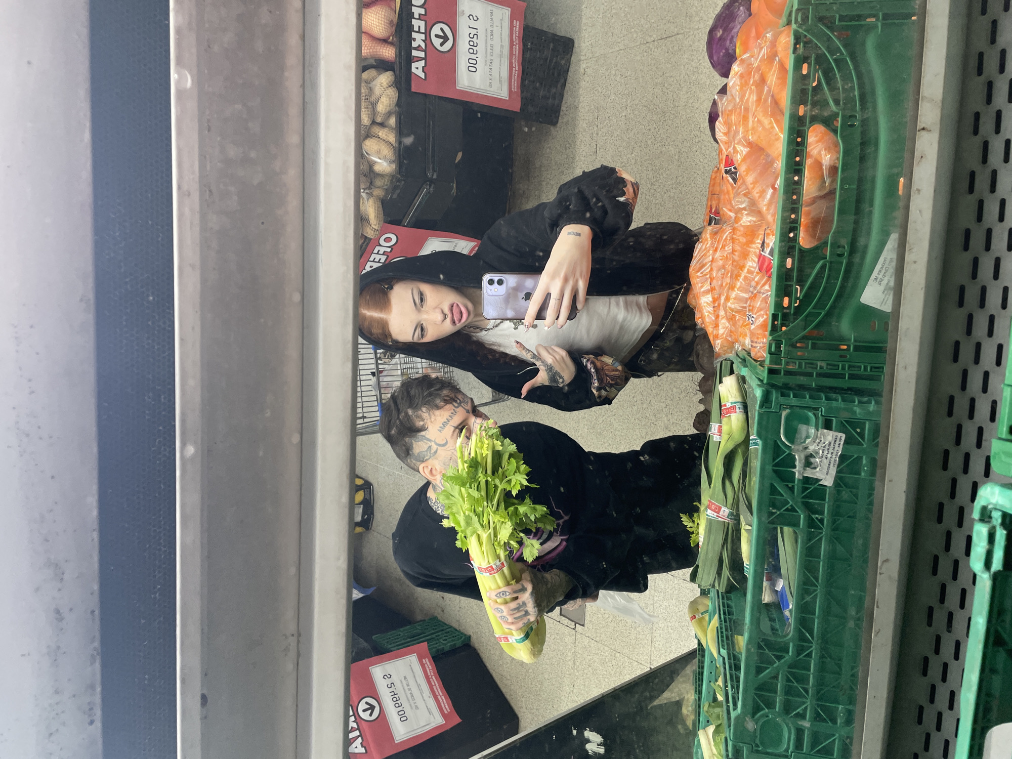 The image depicts a reflection in a supermarket mirror. A woman with red hair is holding a smartphone and taking a selfie. Next to her, a man with face tattoos is holding a bunch of celery. They are standing in the produce section of the supermarket, with crates of carrots, beets, and other vegetables visible. The image is an interior shot of a supermarket, likely in Buenos Aires, Argentina. The reflection shows price tags and signs with writing in Spanish, which is a key indicator of location.