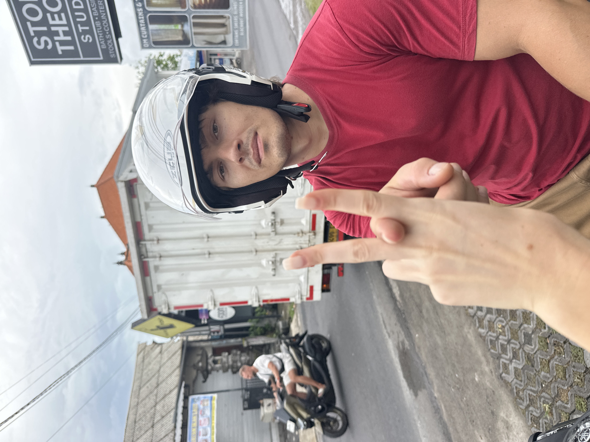 A medium shot, at an oblique angle, depicts a person wearing a white motorcycle helmet and a red shirt, extending their middle finger. The background includes a white delivery truck, a storefront with a sign partially obscured but appearing to say "STONE THE STUDIO", and another motorcycle with a person on it in the street. The surroundings suggest an urban or semi-urban setting, consistent with Munggu, Indonesia. The ground visible is paved with interlocking brick.