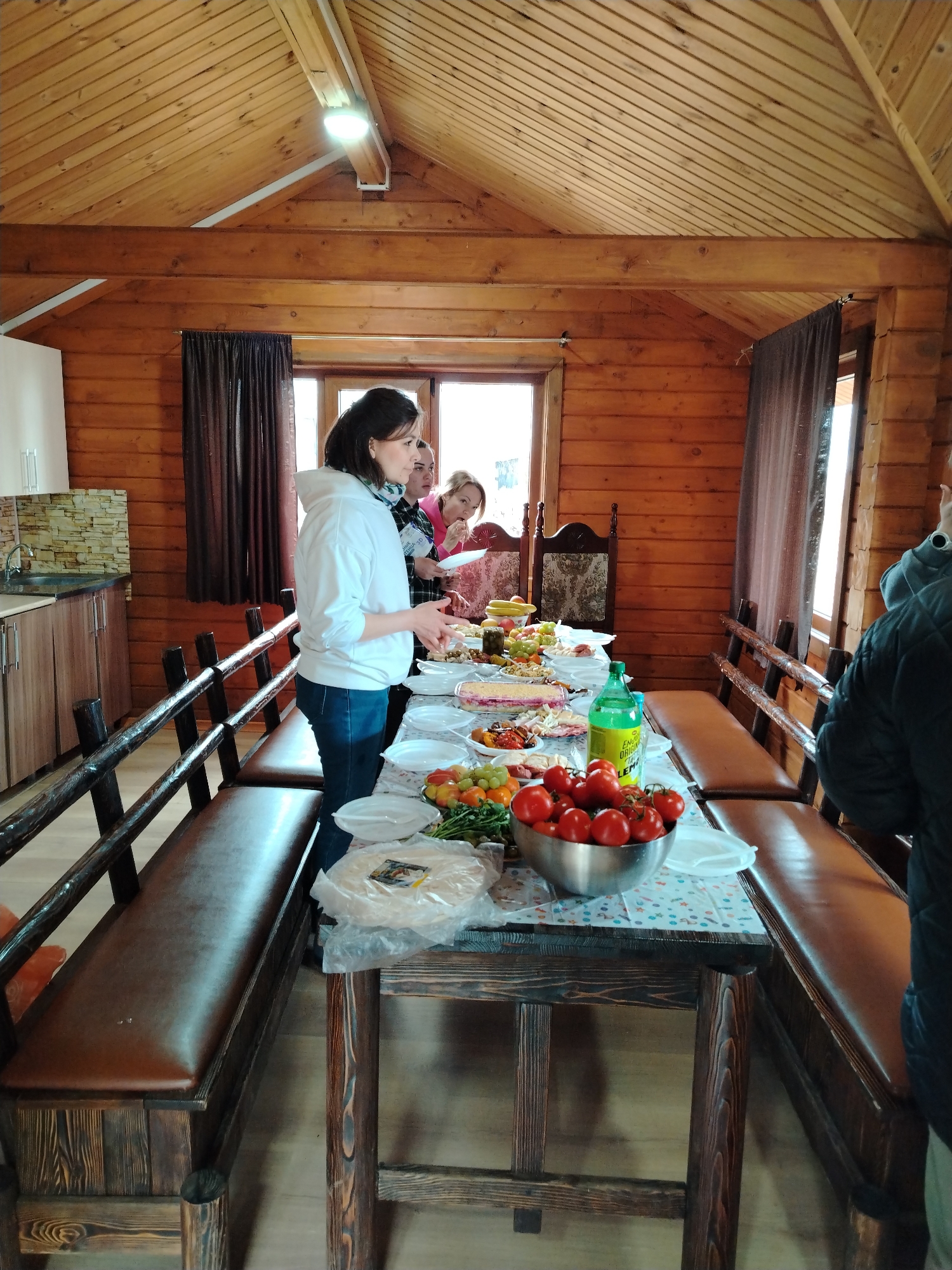 The image shows a group of people gathered around a long table laden with food inside a log cabin. The cabin has light-colored wooden walls and a vaulted ceiling with a modern light fixture. Windows with brown curtains provide natural light.
Several people are visible, with a woman in a white hoodie and jeans standing at the table, possibly serving or organizing the food. Other individuals are behind her, partially obscured, and one person in a dark jacket is near the edge of the frame.
The table is covered with a patterned tablecloth and holds a variety of food items including tomatoes in a metal bowl, various fruits, salads, plates, and a bottle of lemonade. The table is flanked by long benches with dark brown leather cushions and wooden frames. To the left, a small kitchen area with cabinets and a sink can be seen.
Given the information requested, it is implied that the location of this scene is in the city of Chaikovsky, Russia.