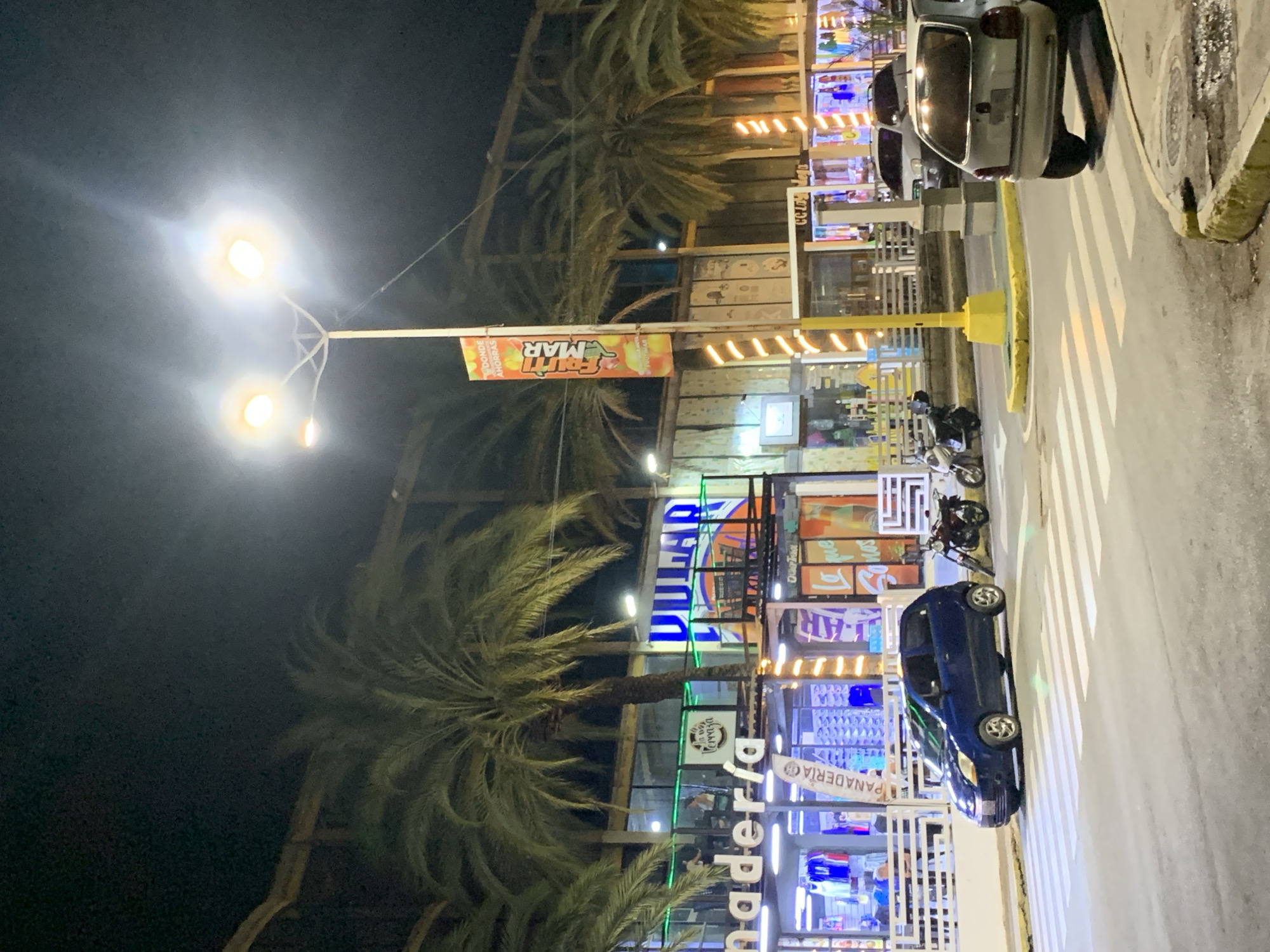 The image depicts a street in Juan Griego, Venezuela at night. Street lights illuminate the scene. Palm trees are visible on the left side of the street. Commercial buildings line the right side of the street. Parked vehicles, including a white car and a blue car, are present. A motorcycle is parked on the side of the road. Business signage in Spanish is visible. A "Frutti Mar" banner is hanging. The "Dulari" and "Panaderia" signs are visible as well.