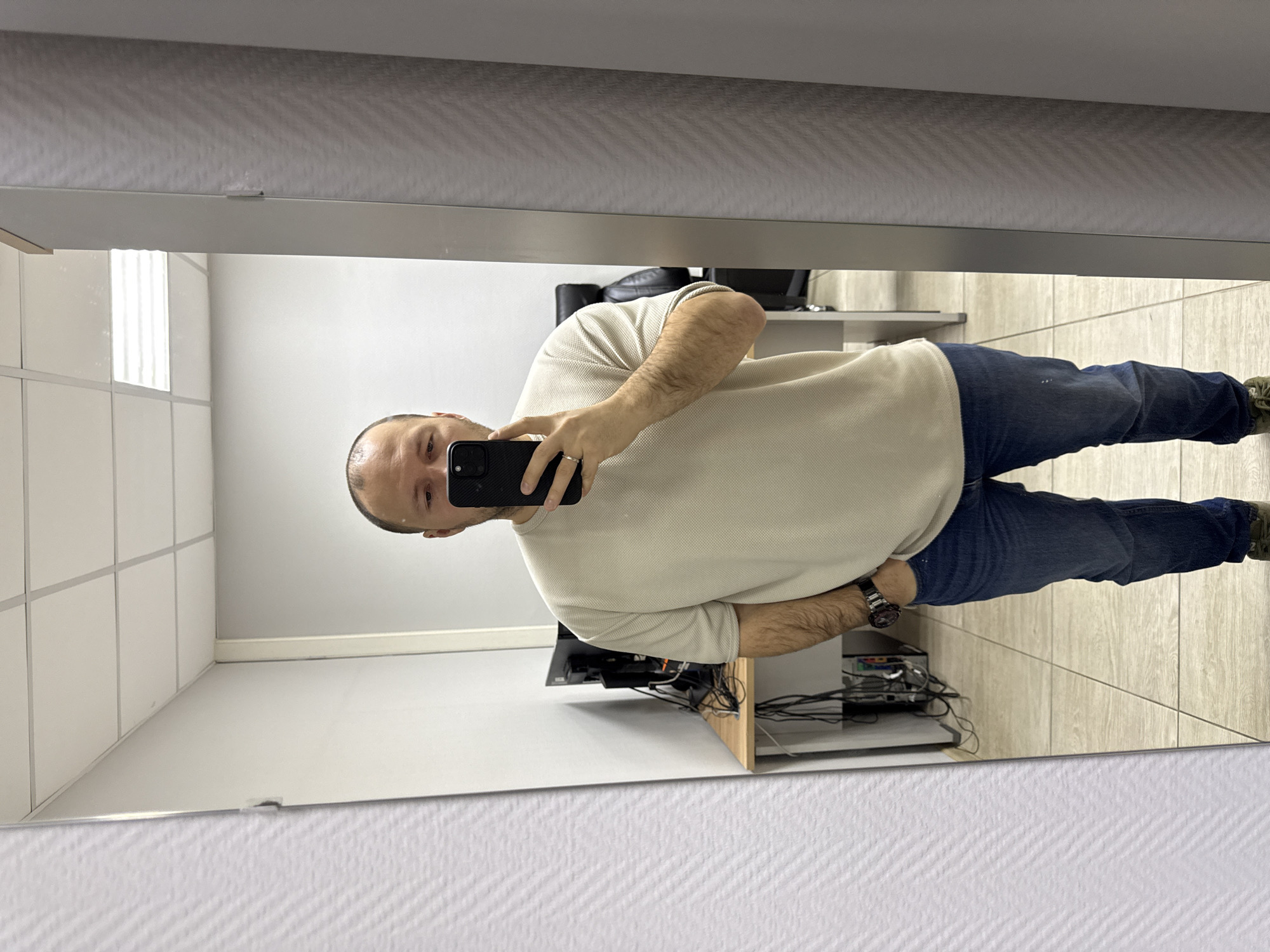 A man is taking a selfie in a mirror. The man is wearing a light beige sweater and blue jeans. He has short, thinning dark hair. He is holding a black smartphone in his left hand, which is visible in the reflection. He has a ring on the fourth finger of his left hand. The mirror is reflecting an office space with a desk cluttered with electronic devices and wires, a chair, and white tiled walls. The floor is tiled with light wood-patterned tiles. The mirror is framed by a white textured wall. The location is likely an office building, but specific details regarding its location in Krasnoyarsk, Russia are not discernible from the image.