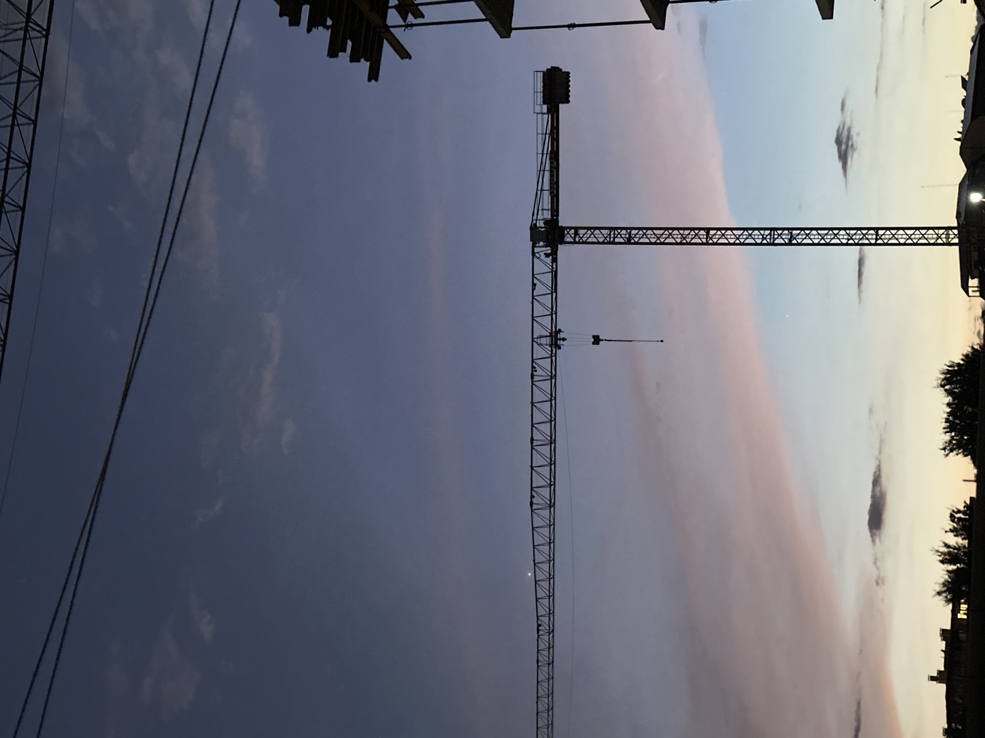 The image shows a construction site at dusk in Sofia, Bulgaria. A large tower crane is the central subject, silhouetted against a sky of deep blue transitioning to lighter shades of pink and orange near the horizon. The crane's boom extends horizontally across the frame, with its lattice structure clearly visible. In the background, the outlines of buildings and trees are visible against the fading light. Two parallel power lines are stretched across the upper portion of the image. The overall atmosphere is one of calm and transition, typical of the end of the day in an urban environment.