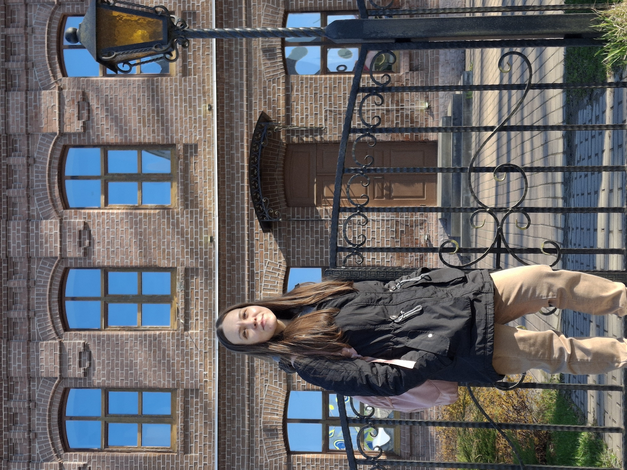 A young woman stands outdoors on a bright, sunny day in Sarapul, Russia, positioned slightly to the right of the frame, behind an ornate black metal fence. She looks directly at the camera with a neutral expression. She is wearing a black jacket with distinctive toggle closures, light brown or tan pants, and has a light pink backpack or bag on her left shoulder.
Behind her is a multi-story building constructed from reddish-brown bricks. The building features several arched windows with reflective blue glass, mirroring the clear sky. A wooden door with an ornate arched canopy is visible towards the center of the building. An old-fashioned, dark metal street lamp with a yellow-tinted glass shade is attached to the brick wall on the upper left.
The foreground shows a paved ground, possibly cobblestones or bricks, with strong shadows cast by the fence, indicating bright sunlight. Some green grass and bushes are visible behind the fence and in the lower right portion of the frame. The black metal fence features intricate scrollwork and decorative elements. No visible text is present in the image.