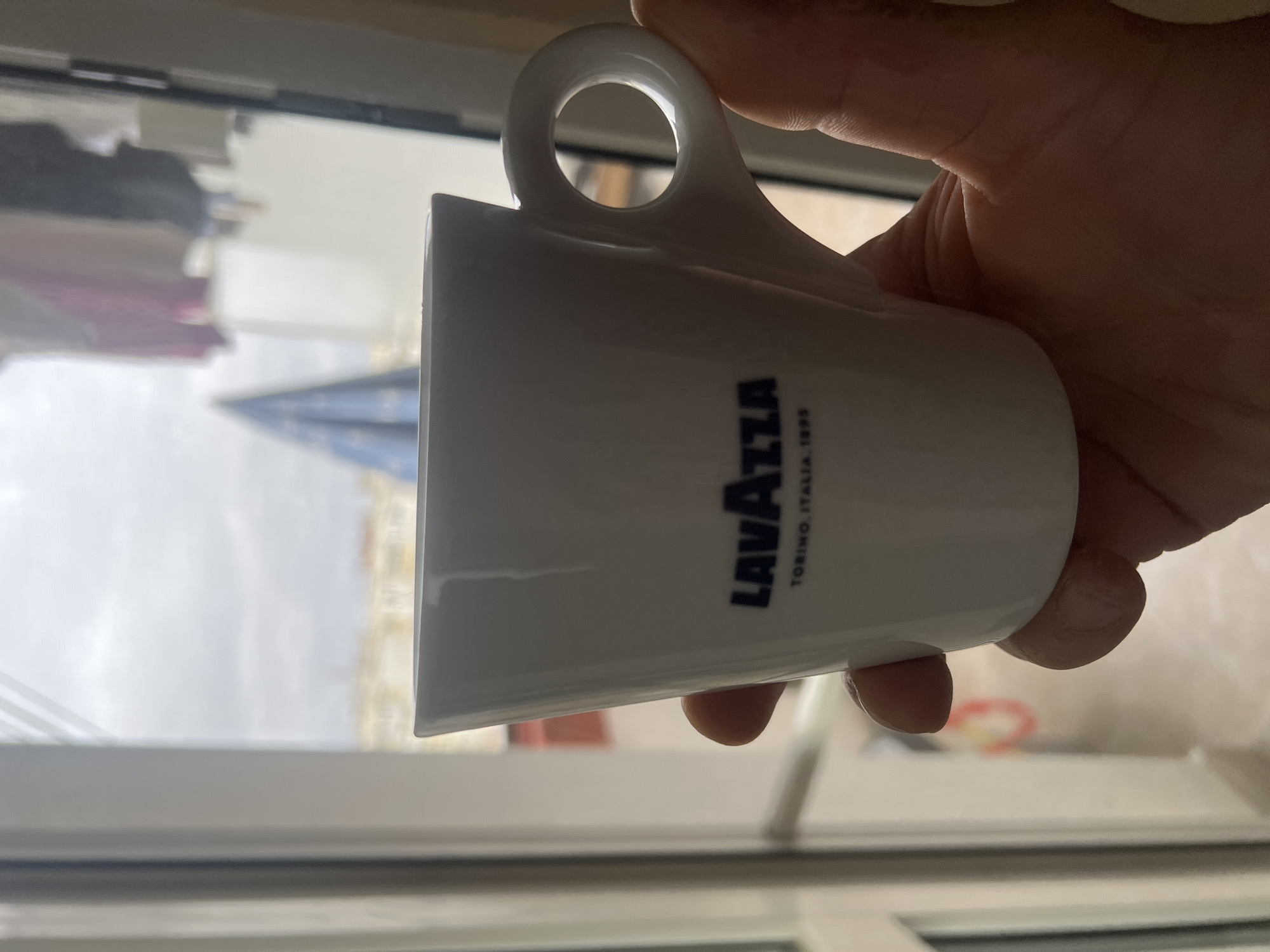 A hand holds a white Lavazza coffee cup with the logo in dark blue, angled slightly to the right. The cup is in front of a window. Outside the window, in soft focus, is a building with a glass or metal pyramid-shaped roof or architectural feature. The scene is in Sofia, Bulgaria, based on architectural style and available geolocation data. The background objects are typical of an urban environment, but precise location cannot be determined without additional visual context.