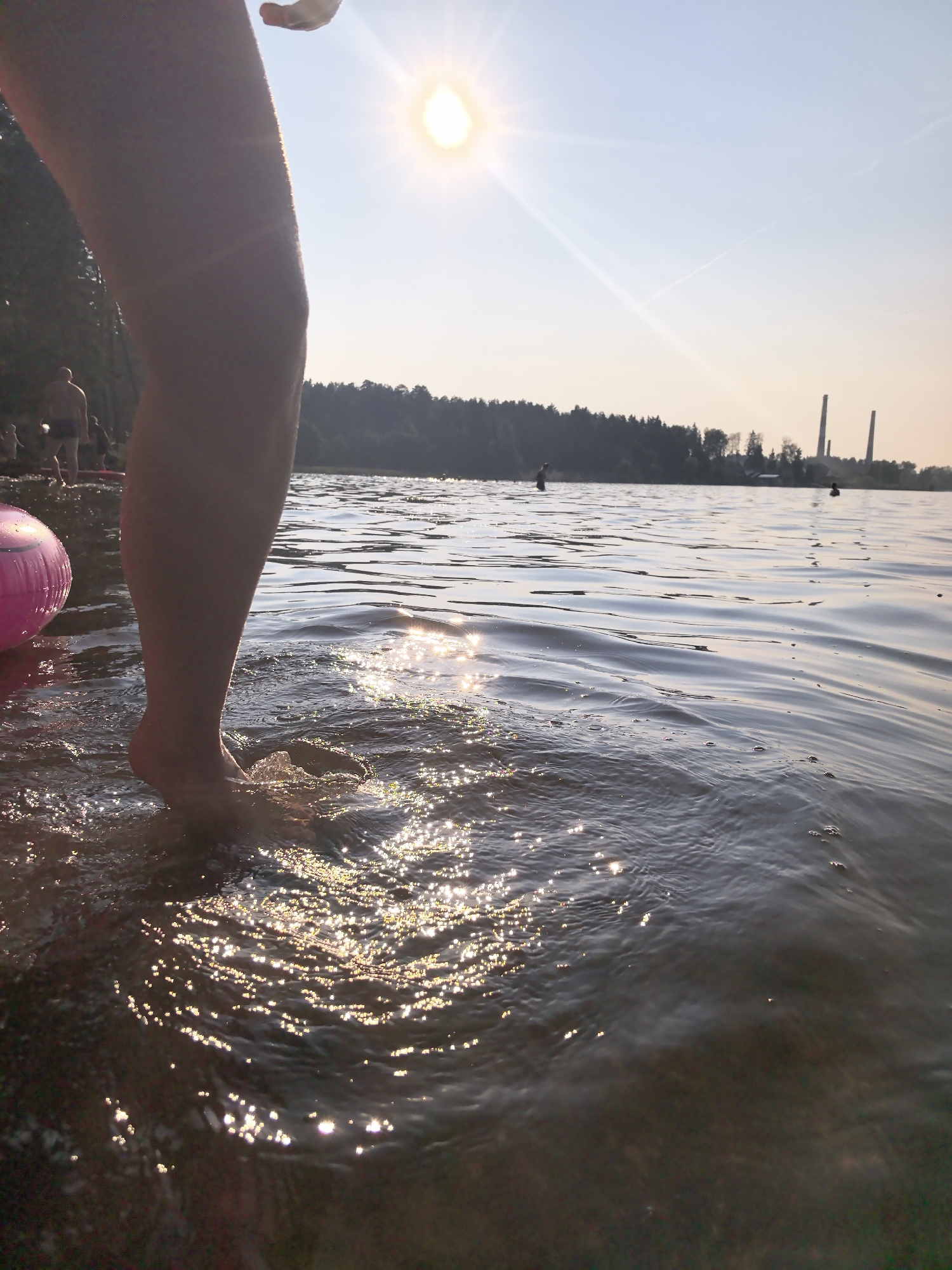A low-angle, medium shot captures a person's bare leg and foot entering the shallow water of a lake. The sun shines brightly in the upper center of the frame, creating sparkling reflections on the water's surface. In the background, a line of trees borders the far side of the lake, with two smokestacks visible above them. The sky is clear with a light haze. Several other individuals are present in the water and on the shore in the distance, appearing to be enjoying recreational activities. A bright pink inflatable ring rests on the shore to the left of the main subject. The scene appears to be set in a natural environment, possibly a beach or recreational area, likely in the vicinity of Chaikovsky, Russia, given the presence of industrial smokestacks in the background which are characteristic of the city's landscape.