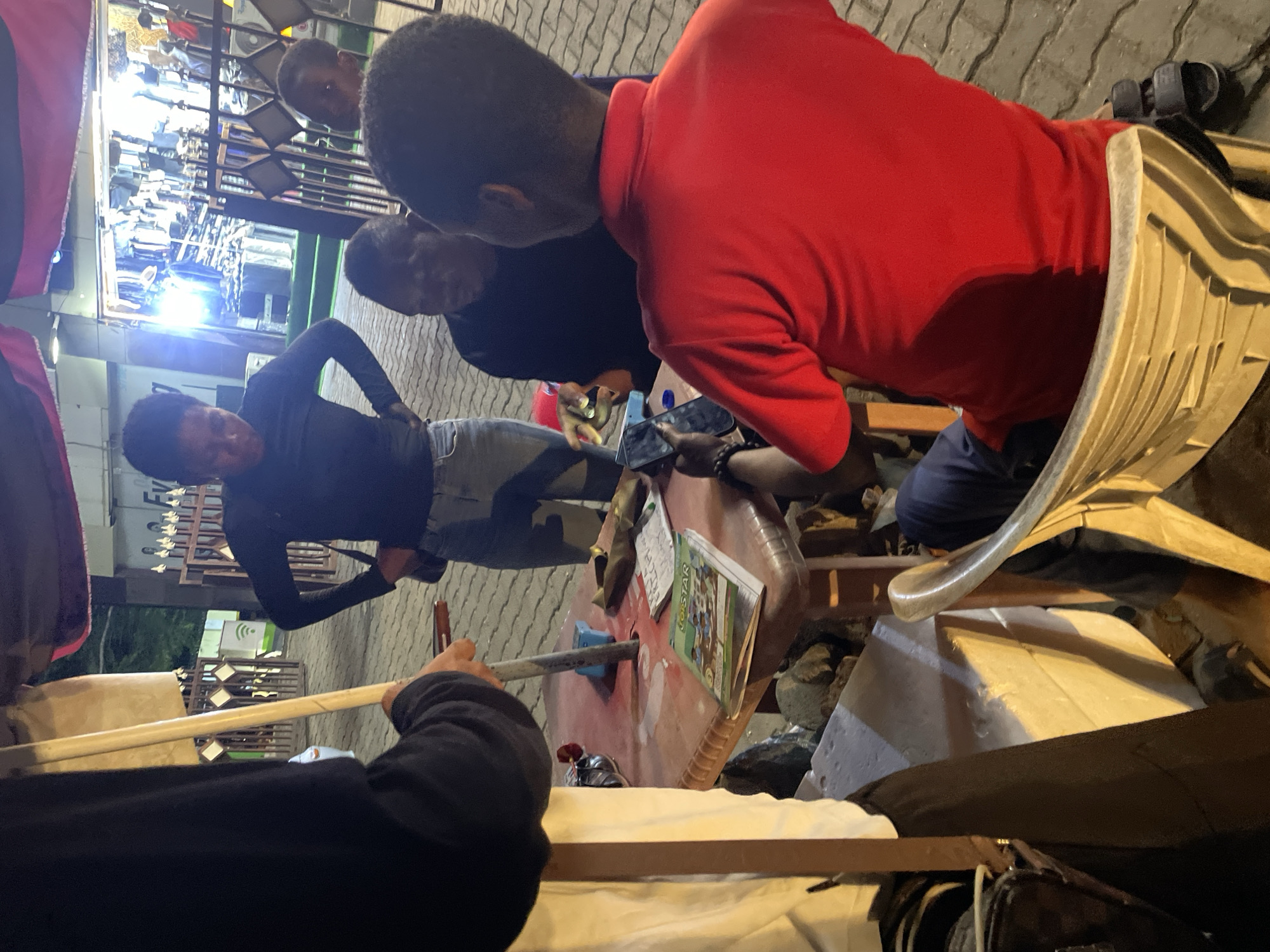 A group of men are gathered around a red table outdoors in Abuja, Nigeria. The table has a book or magazine on it. One man is wearing a red shirt and sitting in a white plastic chair. Another man is holding a cell phone. There is a clothing store in the background. The ground is paved.