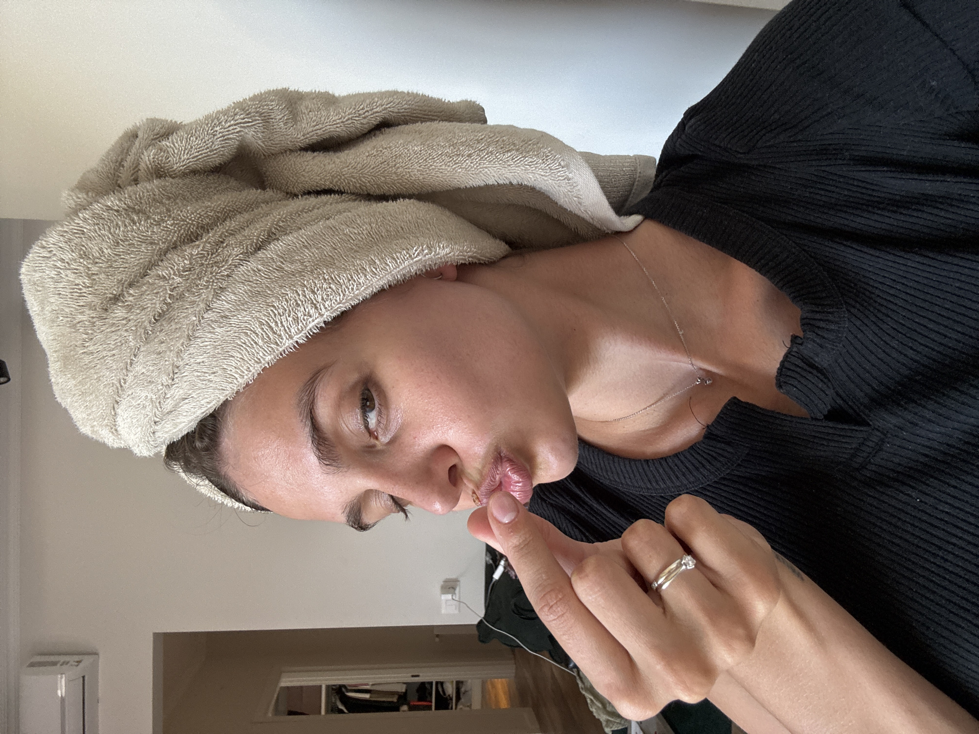 The image captures a person, likely a woman, from the chest up, with her head tilted. She has a light beige terrycloth towel wrapped around her head, suggesting she may have just washed her hair. She is wearing a black ribbed top or dress and a slender silver necklace. Her lips are puckered in a "kiss" or "shhh" gesture, with her right index finger gently touching her upper lip. A silver ring with a small, clear stone is visible on her left ring finger. Her skin appears fresh and dewy. The background is a plain, light-colored wall, possibly white or off-white. On the left side of the background, a white vent or air conditioning unit is partially visible. A hint of another room or doorway can be seen in the lower right background. The setting appears to be an indoor, private space, possibly a home. No text is visible.