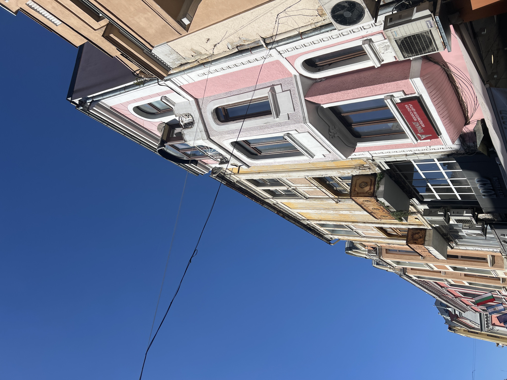 The image shows a street scene in the city of Unknown, Unknown. The perspective is looking up at the buildings, showcasing a variety of architectural styles. The sky is clear and blue. The buildings are varied in color and height, with details such as windows, balconies, air conditioning units, and decorative trim. Some buildings feature awnings or signage. A black electrical wire extends across the frame against the blue sky.