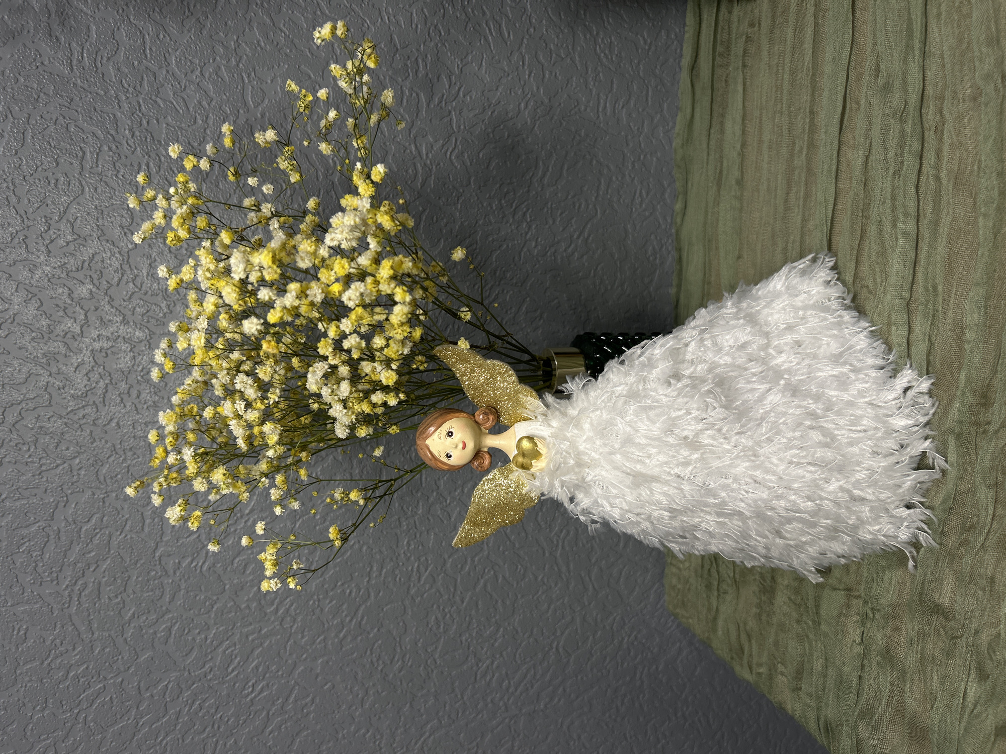 The image presents a still life composition featuring a decorative angel figurine alongside a bouquet of baby's breath flowers, set against a two-part textured background.
The angel figurine is positioned centrally, characterized by a sweet, porcelain-like face with delicate features, including small black eyes, red lips, and light brown, sculpted hair. Its body is adorned with a voluminous, fluffy white dress, reminiscent of soft yarn or feathers. Large, golden, glittery wings protrude from its back, and a small golden heart decorates its chest.
To the angel's left, a dense bouquet of baby's breath (Gypsophila) flowers emerges from a dark, partially visible base, which also seems to hold the angel. The tiny blossoms are a harmonious blend of white and soft yellow, creating a light, airy cluster on their slender green stems.
The background is divided into two distinct textures. The left and upper portion features a dark gray surface with an irregular, embossed, or crinkled texture, possibly wallpaper or a decorative panel. The right side is composed of a vertically draped, light olive-green fabric, distinguished by a fine, crinkled pattern that adds depth and softness to the scene.
The lighting is diffused and even, emphasizing the intricate textures of the angel's dress, the delicate flowers, and the varied surfaces of the background. No specific activity or event is depicted, as the image captures a static decorative arrangement. There is no visible text. The scene is indoors, likely a display on a flat surface. No direct location cues suggest Saint Petersburg, Russia, from the visual information alone.