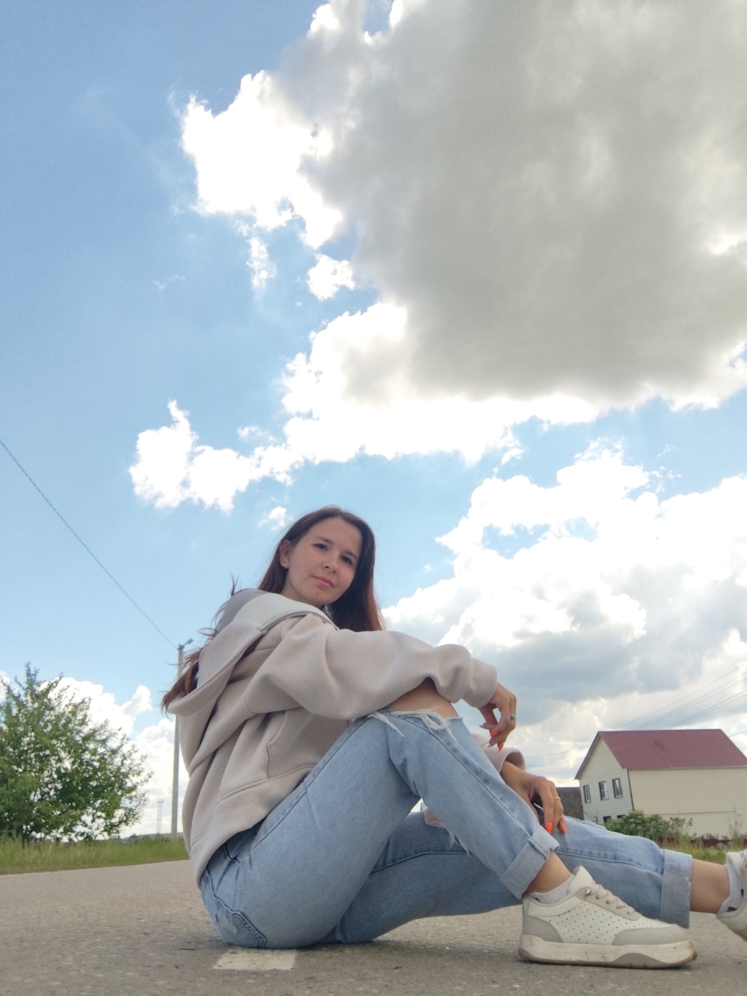 A woman is sitting on an asphalt surface outdoors with a cloudy sky overhead. The woman is wearing a light beige hoodie, light blue jeans with a rip at the knee, and white sneakers. Her hair is long and brown. Behind her, there is a grassy area with a tree on the left and a two-story building with a red roof on the right. Power lines are visible in the sky. The location is Фоки, Russia.