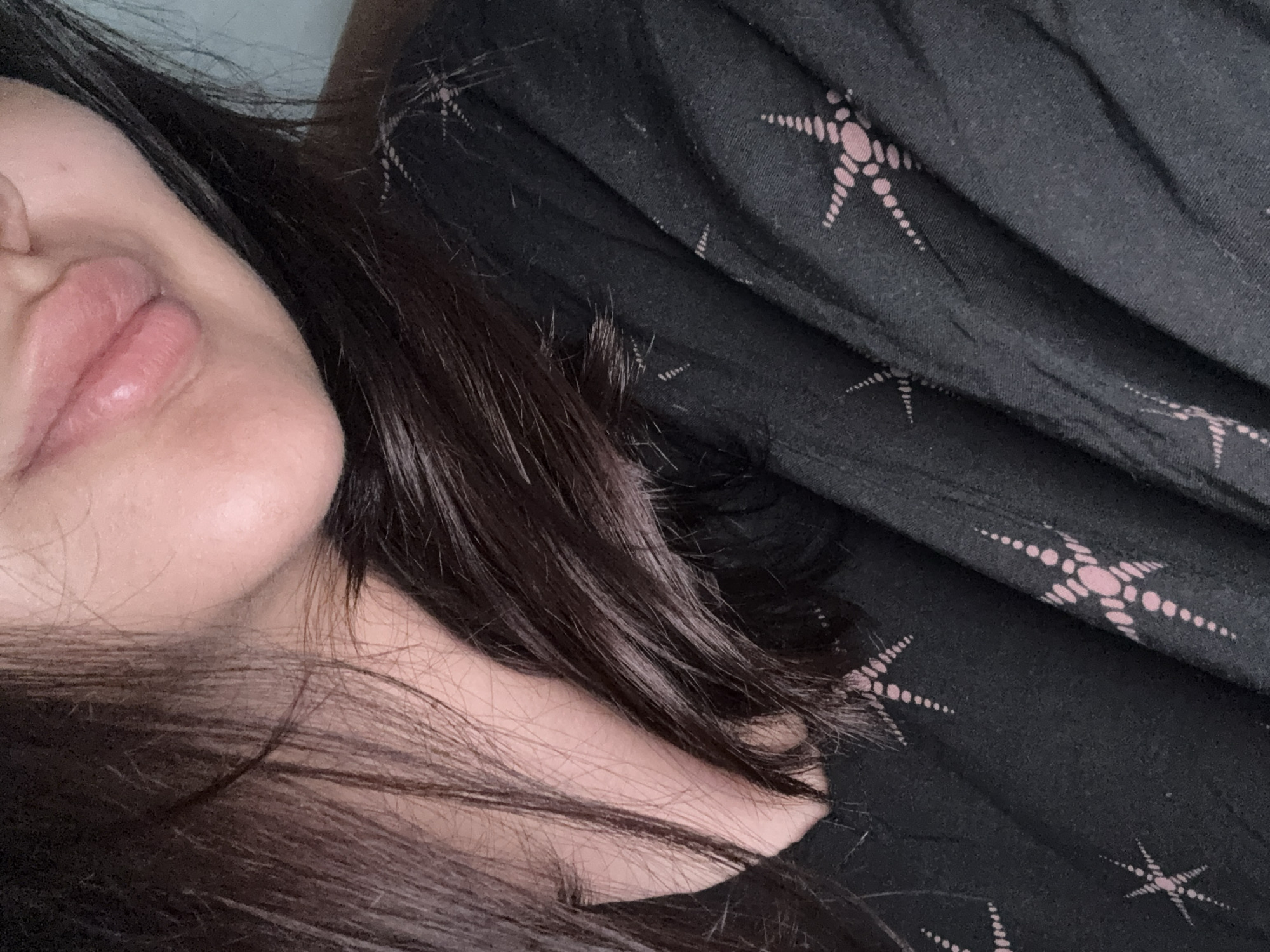 The image shows a close-up of a person's face and chest, with a focus on their lips and dark hair. The person is wearing a black top with a repeating star-like pattern in a light pinkish hue. The background is a plain, light blue. There is no identifiable city or location, including Misaylovo, Russia, visible in the image. The scene is static, with no discernible actions or interactions.