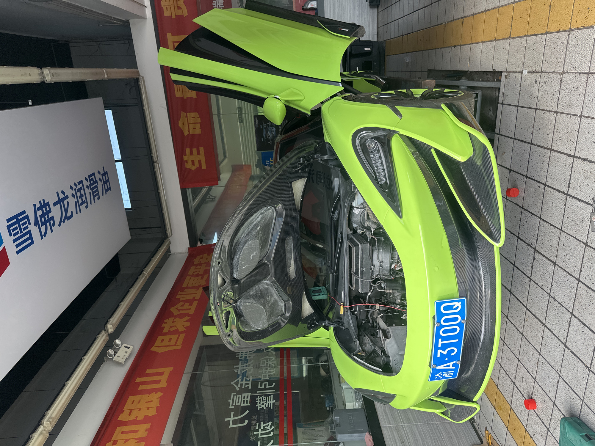A bright green McLaren sports car is parked indoors with its hood and dihedral doors open, revealing its engine compartment. The car is positioned over a hydraulic lift, suggesting it is undergoing maintenance or repair. The license plate on the front of the car reads "A.3TQQQ". The setting appears to be a car repair shop or a garage, with tiled flooring and fluorescent lighting overhead. Banners with Chinese characters are visible in the background, one of which is red and appears to be advertising something related to "life". Another banner, with blue lettering, is positioned above the red one. The environment is clean and organized, consistent with a professional automotive service center. The car itself is the primary focus, its vibrant color and open design making it stand out in the scene.