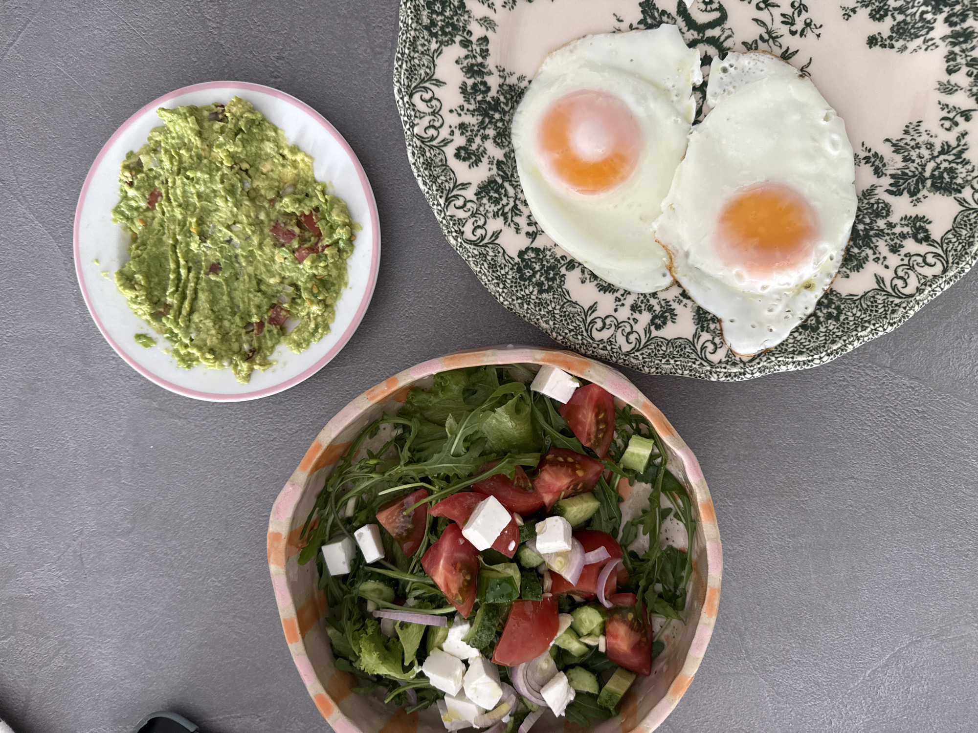 An overhead shot captures a vibrant and diverse meal laid out on a textured, plain gray tabletop, typical of a healthy breakfast or brunch in Tel-Aviv, Israel.
In the upper left, a small white plate with a pink rim holds a generous serving of mashed avocado or guacamole, showing hints of red pieces, likely tomato, and possibly other ingredients mixed in. Its texture appears chunky and freshly prepared.
To the right, on a cream-colored plate adorned with intricate dark green floral and leafy patterns, sit two perfectly cooked sunny-side up fried eggs. Both eggs feature bright, round orange yolks surrounded by firm, white albumen with slightly crispy edges.
In the lower right portion of the frame, a colorful salad is presented in a distinctive bowl with a striped rim in shades of pink and orange. The salad consists of a base of mixed green leaves, including what appears to be arugula. It is generously topped with large chunks of red tomatoes, diced green cucumbers, thin slices of red onion, and several cubes of white feta cheese.
The overall impression is one of a fresh, wholesome, and appealing meal. There is no visible text in the image.