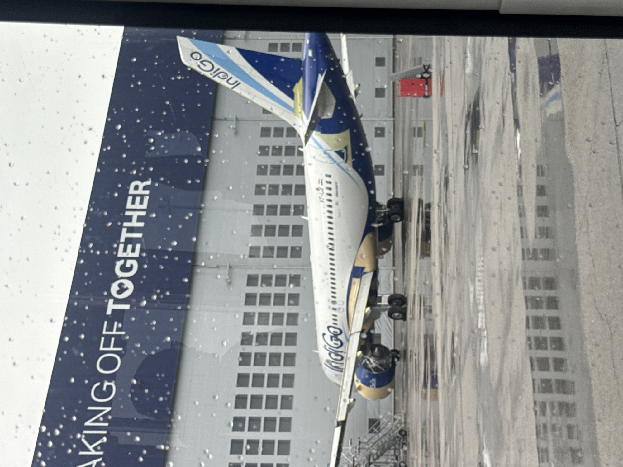 An IndiGo airline passenger jet is seen parked on an airport tarmac, viewed through a window heavily speckled with raindrops and water streaks, indicating rainy weather. The aircraft is predominantly white with distinct blue and gold livery. The "IndiGo" logo is clearly visible on the tailfin and along the fuselage.
On a prominent blue section, likely a winglet or part of the wing, the slogan "TAKING OFF TOGETHER ❤️" is visible. The aircraft's registration, starting with "VT-", can be discerned on the forward fuselage, identifying it as an Indian-registered plane. One of its engines, painted in matching blue and gold, is also partially visible, with ground support equipment like boarding stairs positioned nearby.
In the background, a large, multi-storied grey building with numerous square windows, possibly a hangar or an airport terminal building, stands behind the aircraft. The ground surrounding the plane appears wet. The overall scene suggests an airport environment in Sofia, Bulgaria, during overcast, rainy daytime conditions.