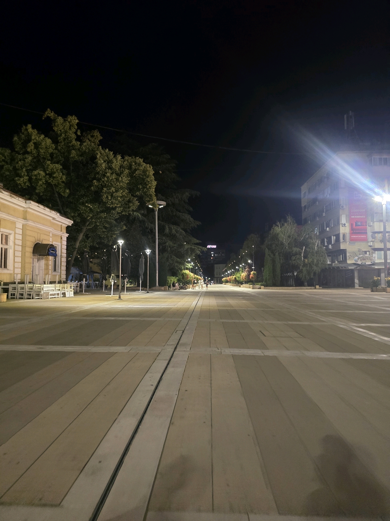 This is a night-time photograph of a pedestrian street in Yambol, Bulgaria. The street is paved with large, light-colored rectangular tiles and features a central channel with metal grating. Lampposts with multiple lights illuminate the walkway. On the left, a yellow building with white tables and chairs in front of it suggests a cafe or restaurant. Mature trees with green foliage line the street, casting shadows. In the background, several multi-story buildings are visible, one of which has a large banner with Cyrillic text and an image. The sky is dark, indicating it is nighttime. A few people are visible in the distance walking along the street. The overall scene conveys a quiet, urban evening atmosphere.