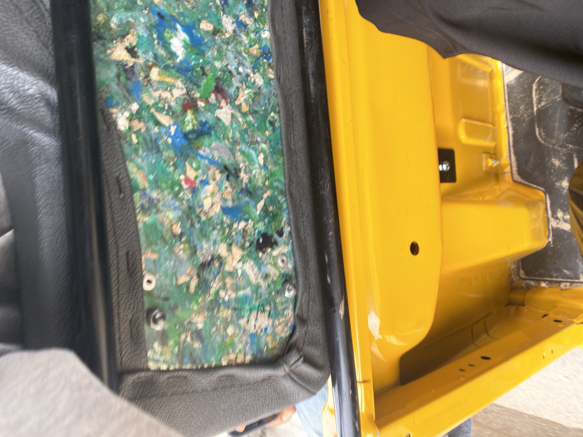 The image shows the interior of a yellow vehicle, possibly a van or a truck. The focus is on the door panel and the surrounding area. The door panel itself is disassembled, revealing layers of soundproofing material, a colorful mix of shredded fibers, possibly recycled materials, in shades of green, blue, and white. This material is held in place by some sort of backing.
To the right of the door panel, the vehicle's interior is painted a bright yellow. There are some structural elements visible, including a recessed area and mounting points for what appear to be fasteners. A black, fabric-like material, possibly clothing, is draped over the top edge of the vehicle's interior.
On the left side of the image, a grey, textured material is visible, which could be part of the vehicle's seat or another interior trim piece. The overall scene suggests some form of repair, modification, or manufacturing process is underway for the vehicle. The lighting appears to be natural daylight, and there are no immediate indicators of the specific time of day or weather. The location context is provided as Jalingo, Nigeria, but there are no distinct visual cues within the image to confirm this specific location. No visible text is present in the image.