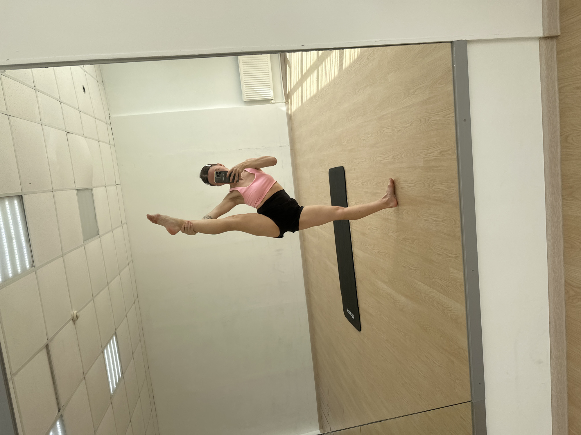 An indoor, rotated mirror selfie captured in Votkinsk, Russia, shows a young woman performing an impressive straddle split, suspended horizontally in mid-air. She wears a pink sports bra/crop top and black shorts, holding a silver/grey smartphone to take the photo with her right hand, while her left hand holds her left ankle, extending her arms to maintain the pose. Her dark hair is pulled back.
The reflection reveals a spacious room, likely a gym or dance studio, with a light-colored wooden floor and a black exercise mat lying flat on it. The mat has the brand name "ZEUS" faintly visible. The walls are mostly white, with a section featuring what appears to be a window with blinds or a ventilation grate, allowing some natural light patterns to fall on the wooden floor. The ceiling, also reflected, is white and features a grid pattern of large, bright rectangular LED lights, providing clear artificial illumination. The mirror itself is framed by a thin grey border. The scene emphasizes extreme flexibility and physical prowess in a well-lit, functional space.