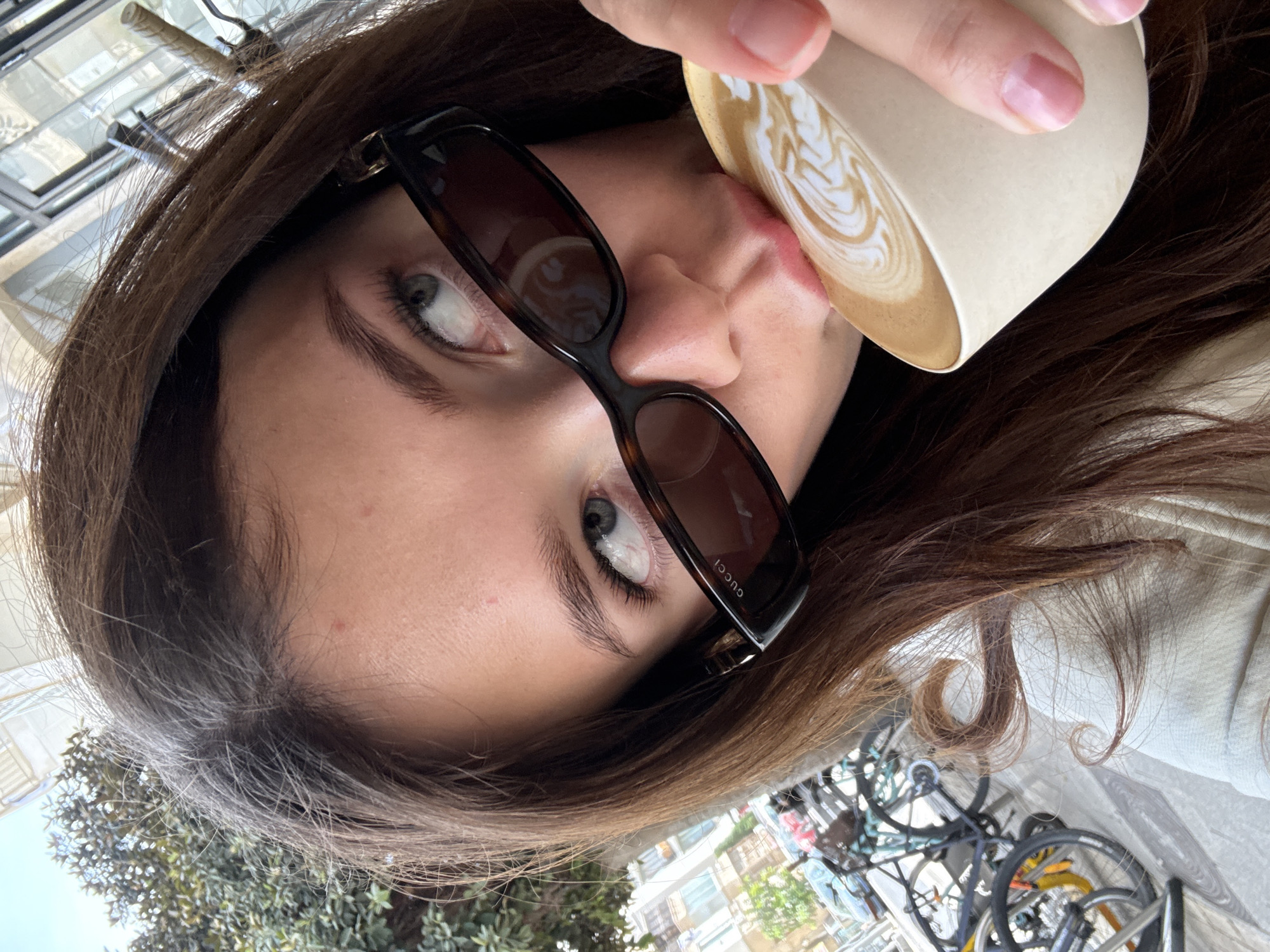 A young woman is drinking from a white paper cup with latte art. She is wearing brown sunglasses with "GUCCI" visible on the side. The woman has brown hair and light-colored eyes. She is likely outdoors, with the background showing trees, buildings, and parked bicycles. The location could be a cafe or restaurant in Tel-Aviv, Israel.