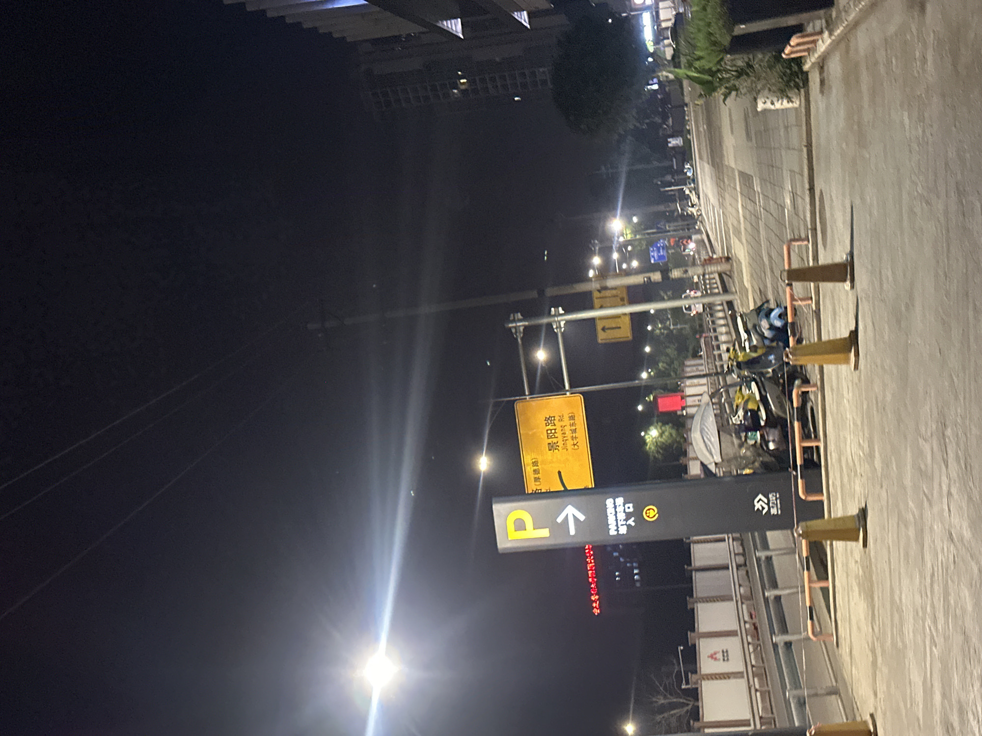 This image depicts a street scene at night in the Chongqing High-tech Zone, China. A tall, dark sign with a yellow "P" and an arrow points to the right, indicating parking. Below this, text reads "PARKING ENTRANCE". To the right of the parking sign is a yellow road sign with Chinese characters and English text reading "Jingyang Rd. (Daqingcheng East Road)". The street is paved with concrete and bordered by a sidewalk with patterned tiles. Several streetlights illuminate the scene. In the background, there are buildings, including a tall, modern high-rise. Parked on the side of the street are several motorcycles and scooters, some covered with tarps. Orange and black striped barriers are visible along the edge of the road. The overall impression is that of a well-lit urban environment at night.