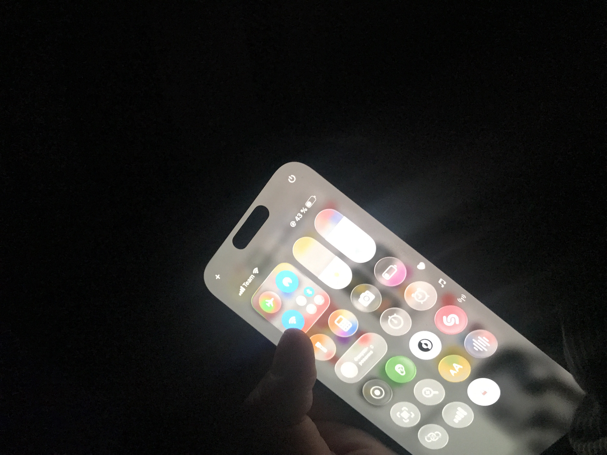 A dimly lit image shows a hand touching the screen of a mobile phone. The phone displays a screen resembling the iOS control center with various circular icons, including Wi-Fi, airplane mode, and brightness control. The phone indicates that it is at 43% battery. The background is primarily dark, suggesting a low-light environment. There's no indication of Yerevan, Armenia, or any specific city or location in the image.