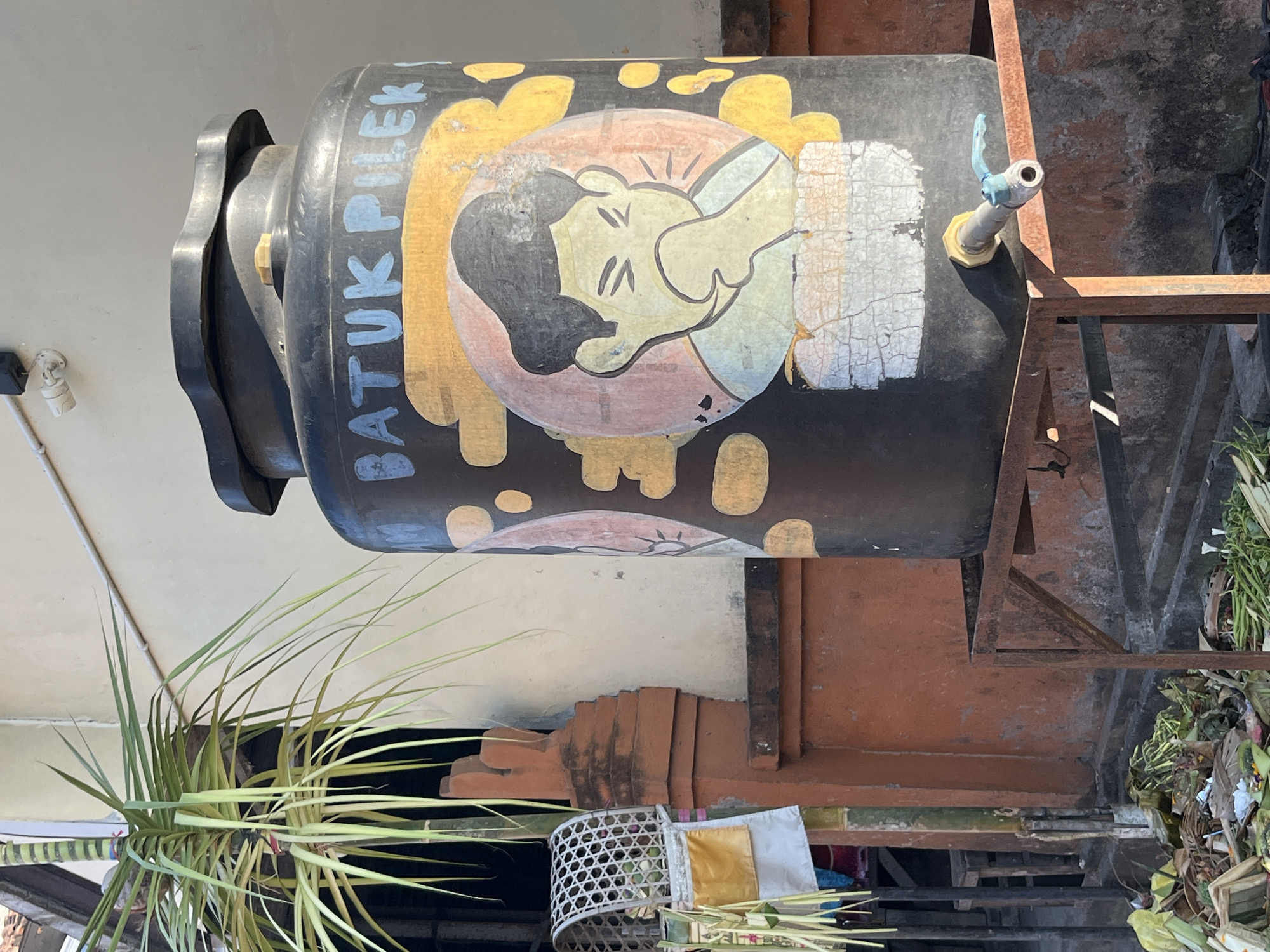 In Sukawati, Indonesia, a large, black water tank sits on a rusty metal stand. The tank features a painted illustration of a person coughing, with the words "BATUK PILEK" written in blue letters above it. A small tap is attached to the lower part of the tank. To the left, a palm-like plant with long, green fronds grows, and further left, a decorative woven basket hangs with some yellow and white fabric. The background shows an orange-brown wall and a cream-colored building. At the base of the tank, there is a pile of discarded organic material, including leaves and green vegetables. A security camera is visible on the cream-colored wall above the tank.