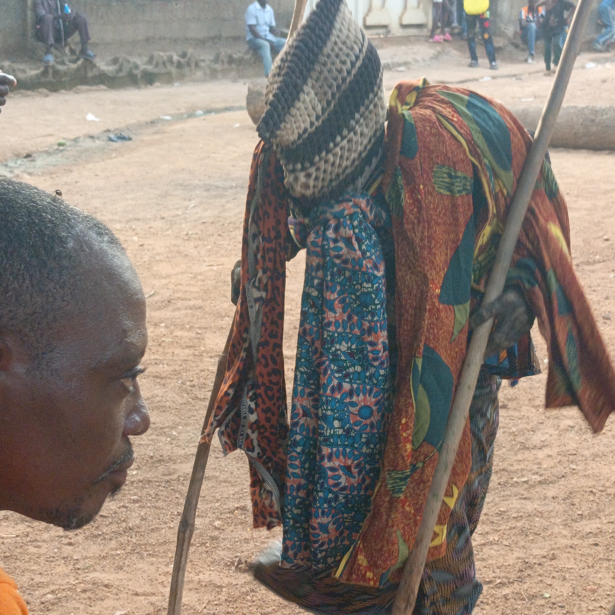 The image shows an African man in Akwanga, Nigeria, looking at a display of fabrics and a knitted hat hanging from wooden poles. The background reveals a public outdoor setting with several people present. The ground is a mix of dirt and sand. In the background, other people are visible, including someone sitting on a ledge and others standing and walking. The overall scene appears to capture a local marketplace or gathering in Akwanga. A fly can be seen on the man's head.