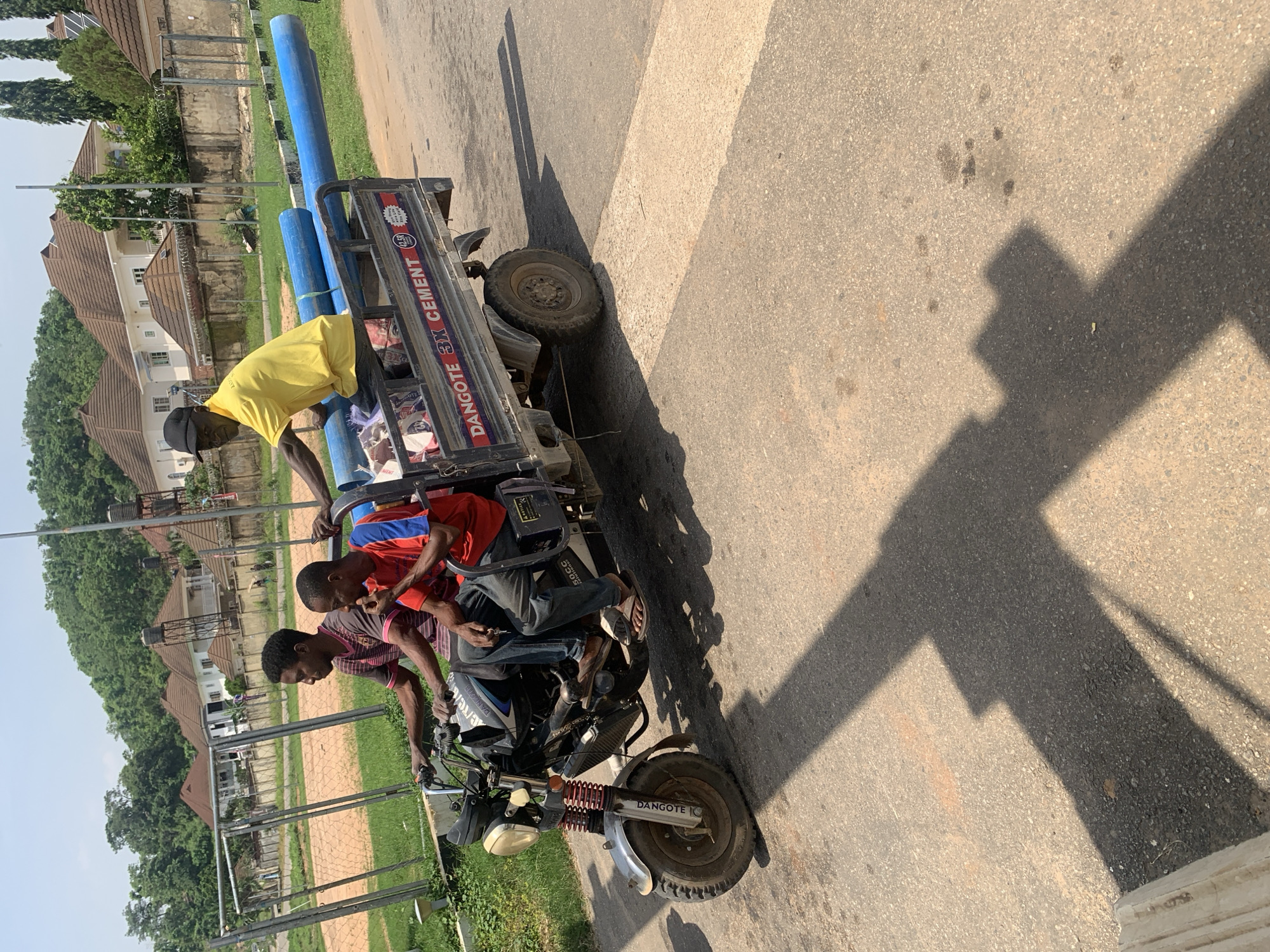 The image captures a scene on a sunny day in Abuja, Nigeria. A man in a yellow shirt and black cap stands on a motorcycle with a trailer attached, holding onto some large blue pipes. Three other individuals are seated on the trailer. Two of them are young men, one wearing a red and blue striped shirt and the other a pink and gray striped shirt. The third person is partially obscured. The trailer is loaded with these blue pipes and some bags, with the branding "DANGOTE 3X CEMENT" visible on its side. The motorcycle itself also has "DANGOTE" written on its front fender. The setting appears to be a roadside, with a paved surface and a grassy area beside it. In the background, there are buildings with brown tiled roofs and trees. The sun casts a long shadow across the road, indicating it is likely midday. The overall atmosphere seems to be one of work or transport.
