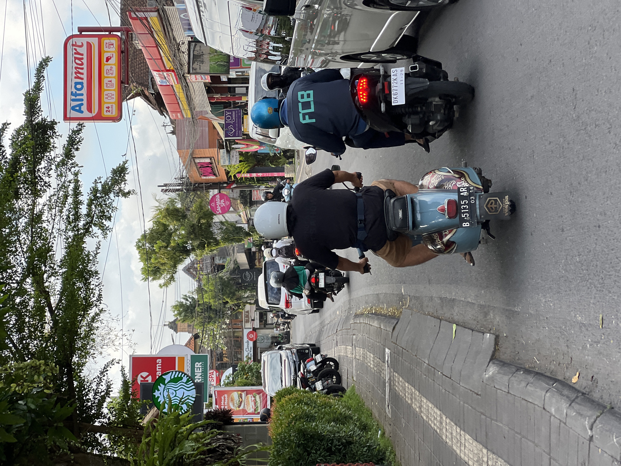 The image captures a street scene in the Ubud District, Indonesia, recognizable by the "Alfamart" store sign on the left side of the frame, along with other businesses such as Starbucks. The focus is on two men riding motorbikes. The first rider wears a black shirt and a white helmet, astride a light blue Vespa scooter, displaying a license plate reading "B 5135 AR" with an expiration date of 03-14. The second rider, behind the first, dons a blue helmet and a dark jacket with the letters "FCB" on the back, riding a motorbike with the license plate "DK6772KAS", expiring 11-29. Both are traveling in the same direction. The image also shows other vehicles and motorbikes in the background, with various shops and buildings lining the street, indicating a busy urban setting.