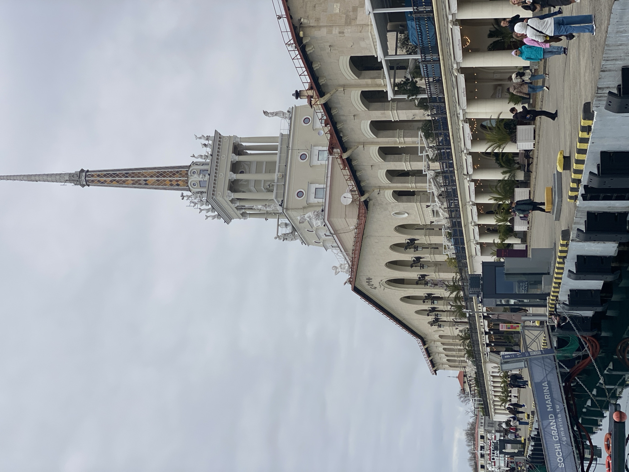 The image displays the Sochi Seaport Terminal building and its adjacent marina in Sochi, Krasnodar Krai, Russia, under an overcast sky. The prominent multi-story structure, primarily of light-colored stone with a red-tiled roof, features a central tower capped by a tall, slender spire. The spire incorporates patterned diamond-shaped panels in yellow, brown, and white hues. The tower itself has multiple levels, including a section with classical columns and decorative statues, specifically white dolphin figures on the top tier and other human-like figures on lower levels. The main building facade is characterized by a series of arched windows on the upper level and a colonnaded lower level, which houses various storefronts and restaurants with outdoor seating areas, awnings, and potted palm trees. The Cyrillic text "СОЧИ" is visible on the upper portion of the building.
In the foreground, the dark water of the marina reflects the muted sky and parts of the architecture. A grey metal gangway, clearly labeled "SOCHI GRAND MARINA sgmarina.ru," extends from the paved pier towards the water. The pier itself features black and yellow striped bumpers, dark mooring cleats, and other marine equipment; red life rings and orange buoys are visible in the water. Approximately 20-30 individuals are present on the paved promenade, casually dressed in jackets and coats, indicating cooler weather. They are observed walking or standing independently, with one person in the mid-foreground looking down at a mobile phone. Dark grey security barriers or structures and additional bollards are situated along the pedestrian pathway.
