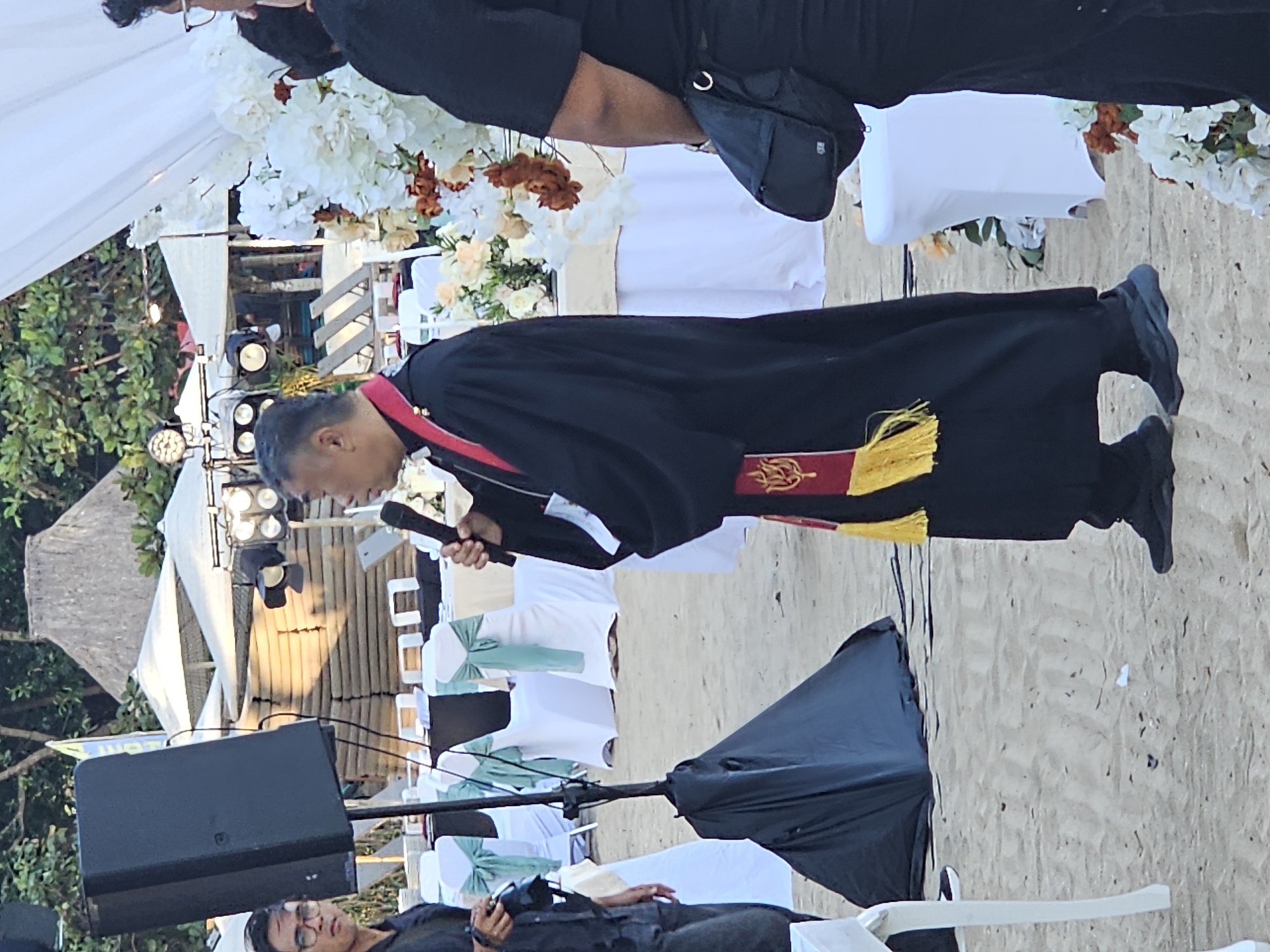 The image depicts an outdoor event, likely a wedding ceremony, set on a sandy beach, possibly in Denpasar, Indonesia. A man, dressed in what appears to be ceremonial or clerical attire, stands center frame holding a microphone. He is wearing a black robe with a red and gold sash draped across his chest, adorned with tassels. His expression suggests he is speaking or officiating.
In the background, a decorated archway with white and cream flowers is visible, suggesting a ceremonial focal point. White chairs, some with mint green sashes, are arranged on the sand, indicating seating for guests. A large black speaker is positioned to the left of the frame. Above the speaker, stage lighting is mounted on a structure that could be part of a temporary setup. A white canopy is also present, offering shade.
The lighting suggests it is daytime, likely late afternoon due to the warm tones of the light. The presence of a beach and the tropical foliage visible on the left side of the image strongly point towards a coastal location. While no text is explicitly visible, the overall scene conveys a formal yet relaxed atmosphere suitable for an outdoor celebration. The people in the image are not clearly defined due to the resolution, but the focus is on the central figure and the event setup.