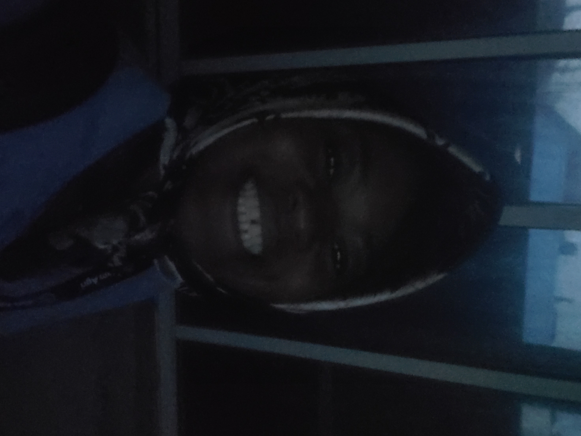 A dark-skinned individual, appearing to be female, is centered in a dimly lit, possibly indoor, environment, purportedly in Kakau, Nigeria. The individual is smiling broadly, exposing white teeth, and looking directly at the viewer. A patterned headscarf, primarily light in color against a darker base, covers the head and frames the face. A segment of a blue garment is visible on the upper torso. On the right side of the individual's chest, a dark lanyard or strap with partial white text, possibly including "Agri", is visible. The background consists of vertical and horizontal structural elements, suggestive of window frames or architectural supports. Beyond these structures, the exterior environment is dark, blurred, and indistinct, with faint blue-grey tones, indicating very low ambient light, consistent with twilight or nighttime conditions.