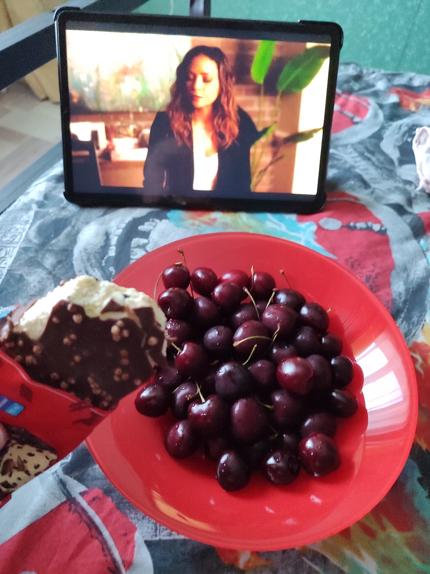 In an indoor setting, a tablet displays a video of a woman with long, dark, wavy hair, wearing a white top and a dark jacket. In the foreground, a red plate holds a pile of dark red cherries, some with stems attached. To the left of the plate, a portion of a chocolate-covered ice cream bar with white cream and puffed rice is visible. The subjects are resting on a colorful, patterned blanket. The location is unspecified within a city.