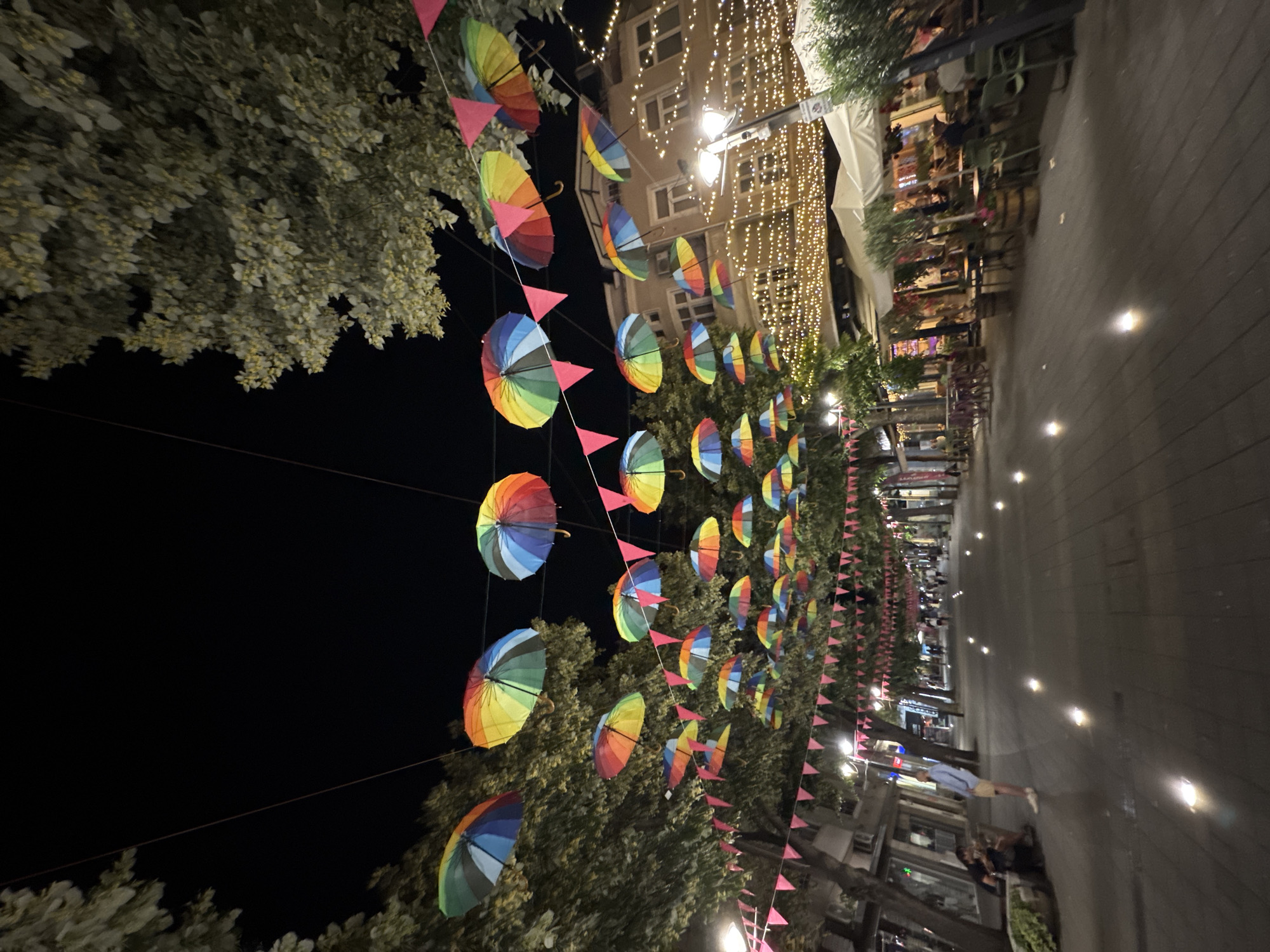 The image depicts a street scene at night, likely in Burgas, Bulgaria, decorated with colorful umbrellas and string lights. The street is paved and lined with trees and buildings, which appear to house shops and restaurants. Several people are visible walking along the street or seated at outdoor tables, suggesting a lively evening atmosphere. The umbrellas, suspended overhead, create a vibrant canopy, with some displaying rainbow patterns. Pink triangular flags are strung along the same wires as the umbrellas, adding to the festive decoration. The lighting from the string lights and ground-level illumination creates a warm and inviting ambiance. The overall impression is of a charming and well-decorated urban area.