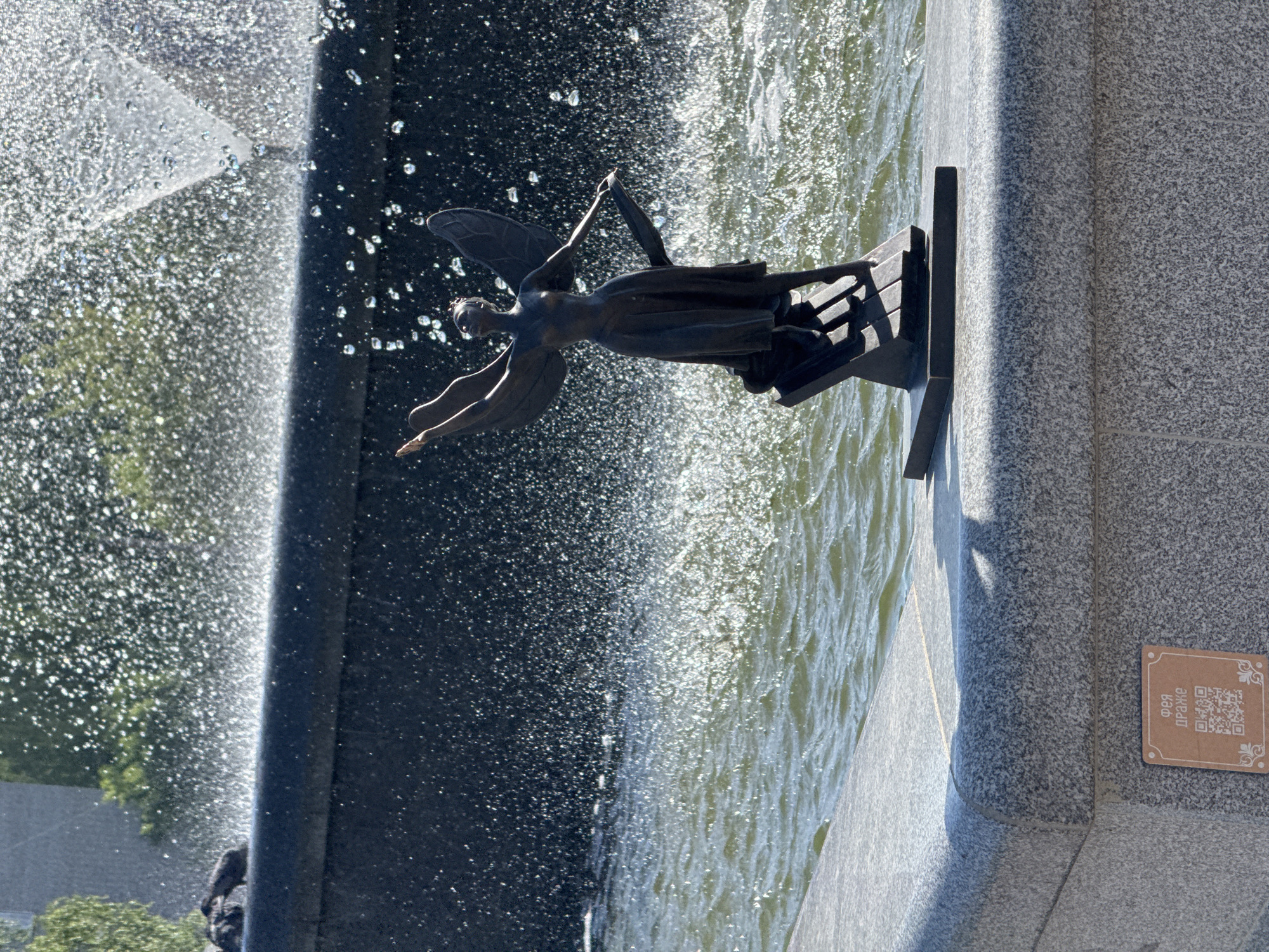 Here's a detailed description of the image:
**Scene:** An outdoor view featuring a statue of a winged figure (possibly an angel or fairy) mounted on a black platform within a fountain. The statue is positioned against a backdrop of water cascading from the fountain and a stone structure. A QR code sign is affixed to the side of the structure.
**Objects:**
* **Statue:** A dark-colored, likely bronze or metal, statue of a winged human figure with outstretched arms.
* **Platform:** The statue stands atop a black platform, possibly made of metal or stone.
* **Fountain:** A fountain structure creating a cascade of water in the background.
* **Stone Structure:** A large, grey stone structure forms part of the fountain's base.
* **QR Code Sign:** A small, rectangular sign with a QR code and text in a foreign language.
**Environment:**
* The environment is outdoors, indicated by the presence of natural light and possibly distant foliage.
* The setting appears to be urban, potentially a park or public square.
**Location**
The statue is the Fairy fountain in the city of Chaikovsky, Russia.
