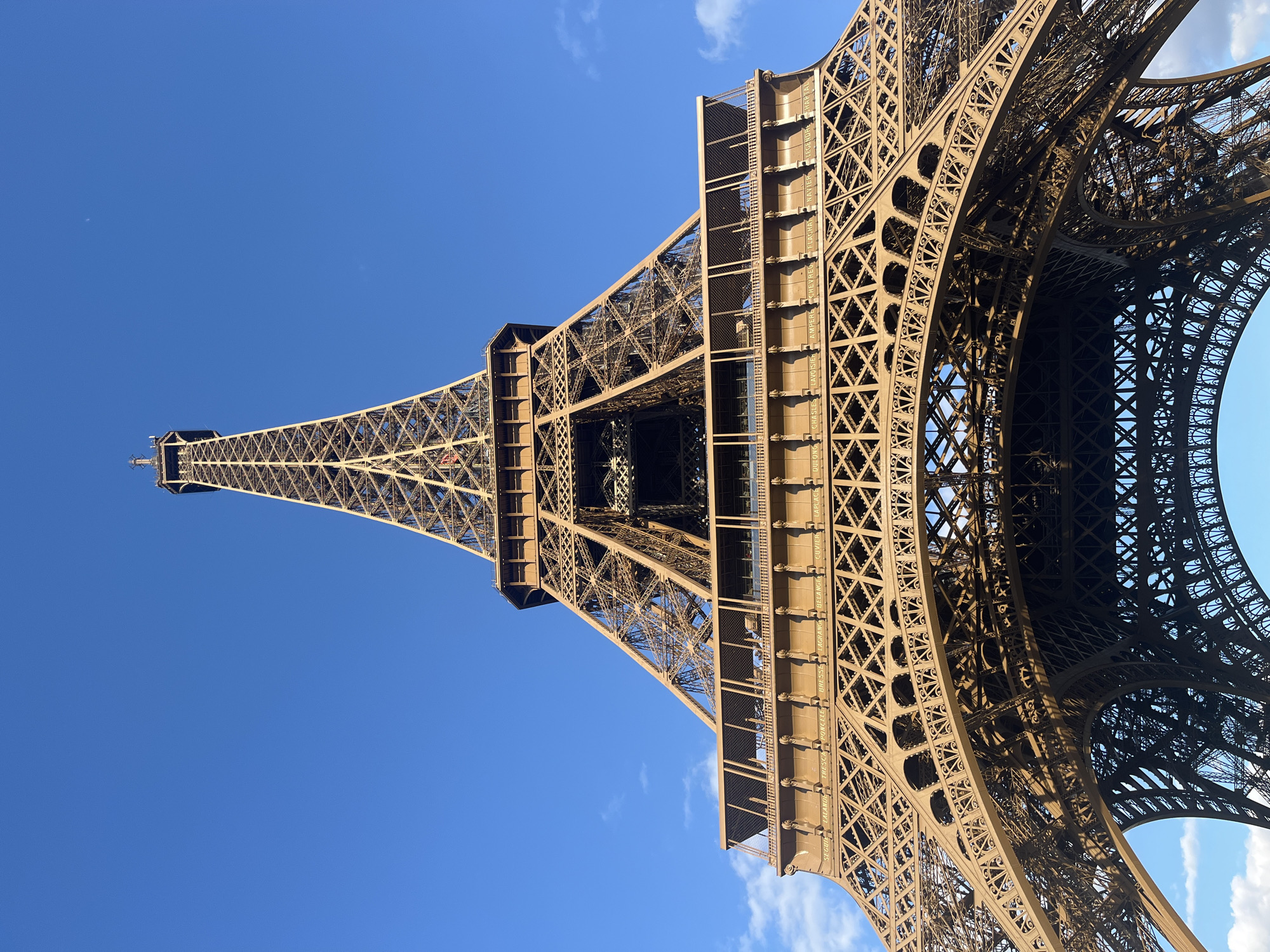 The image shows a low-angle, close-up view of the Eiffel Tower in Paris, France, against a clear blue sky with a few wispy clouds. The iconic wrought-iron lattice structure of the tower dominates the frame, with its intricate geometric patterns and massive scale evident. The lighting suggests it is daytime, likely midday or afternoon, given the bright sunlight casting shadows across the metalwork.
The perspective emphasizes the sheer height and engineering marvel of the tower, with the viewer looking upwards into its structure. The metallic brown color of the tower is prominent, highlighting its weathered texture.
Visible text is inscribed vertically on some of the horizontal support beams of the tower. These inscriptions appear to be names, possibly of scientists or engineers associated with the tower's construction or historical figures. Some legible names include "DULONG," "LAGRANGE," "LAPLACE," "CHASLES," "LEVERRIER," "BAILLY," "DE LA TOUR," and "POINCELET," among others. These names are part of the historical inscriptions on the Eiffel Tower.
The image captures the grandeur and architectural significance of the Eiffel Tower, conveying a sense of awe and wonder. No people are visible in the frame, and the focus is solely on the structure and the sky.