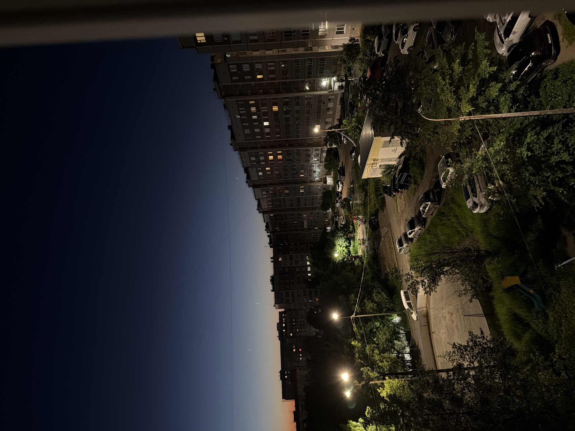 The image depicts a twilight view of a residential area in Izhevsk, Russia. A long, multi-story apartment building dominates the right side of the frame, its windows lit from within. In front of the building, a street is lined with parked cars. Trees and greenery are scattered throughout the scene, with some streetlights illuminating the pathways and parking areas. The sky is a deep blue, transitioning to a lighter hue near the horizon, suggesting the last moments of daylight. In the distance, two crescent moons are visible in the sky. A small playground with a slide is partially visible in the lower left portion of the image. The overall atmosphere is quiet and nocturnal.