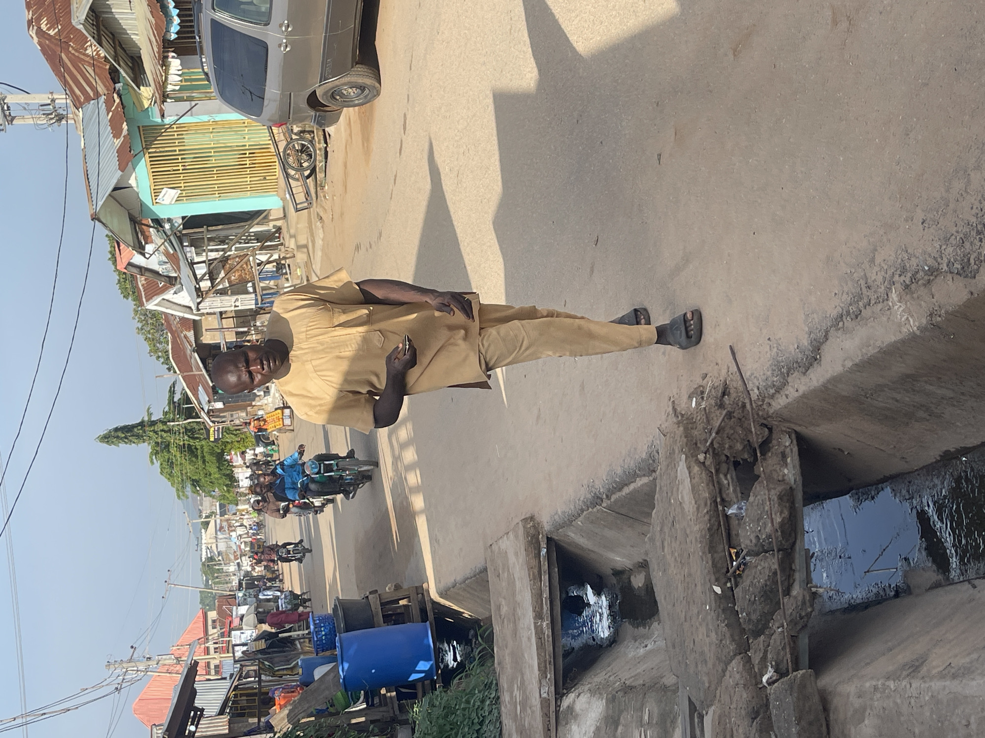 The image captures a bustling street scene in what appears to be Abuja, Nigeria, under bright daylight. In the foreground, a dark-skinned man stands prominently, wearing a light brown or tan traditional African tunic and trousers, with black sandals. He holds a mobile phone in his right hand and looks directly at the viewer with a neutral expression. His shadow extends behind him on the paved road.
To the man's right, a concrete drainage ditch runs parallel to the street, containing stagnant water and some debris. One section of the ditch shows crumbling concrete and exposed rebar. Further down, blue plastic barrels are visible near the drainage.
The street itself is paved but shows signs of wear. A silver minivan is parked on the right, and various motorcycles, known locally as "okadas," are visible in the background, some with multiple riders, indicating active traffic. Buildings line both sides of the street, featuring corrugated metal roofs, and some with brightly colored walls or grilles, such as light blue and yellow. Utility poles with overhead wires crisscross the scene, and some greenery, including a tall, slender tree, can be seen in the distance. The clear sky and strong shadows suggest a sunny day, likely midday or early afternoon. No discernible text is visible on signs or vehicles. The overall atmosphere is that of a lively urban or semi-urban commercial street.